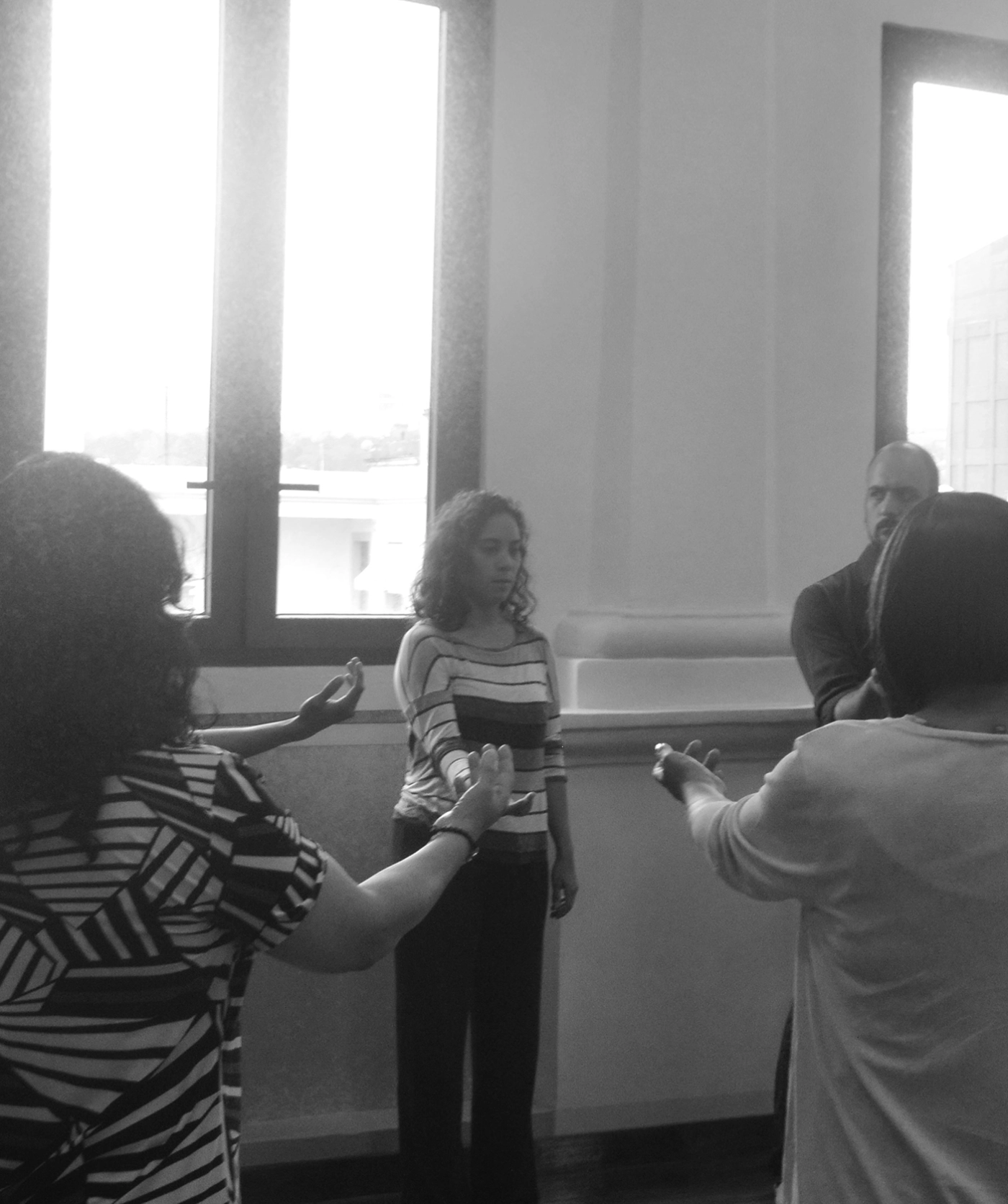Un grupo de personas en una sala, con una mujer de pie frente a los demás, parece participar en una actividad de grupo. La luz entra a través de grandes ventanas detrás de ellos.