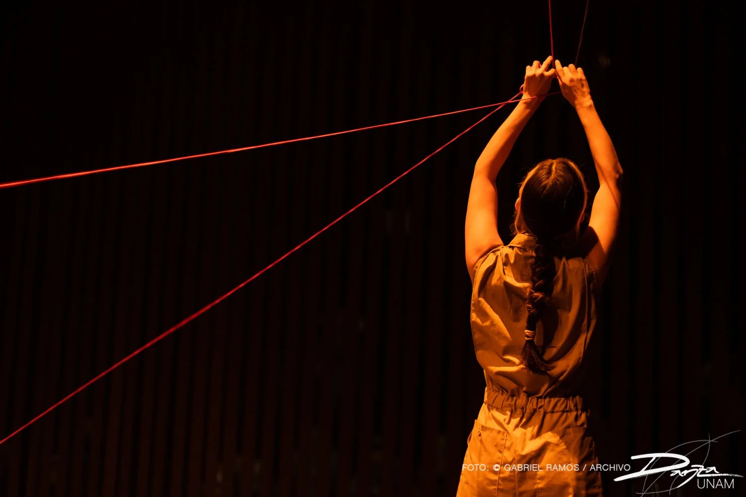 Persona de pie con el cabello trenzado, sosteniendo cuerdas rojas en un entorno oscuro.