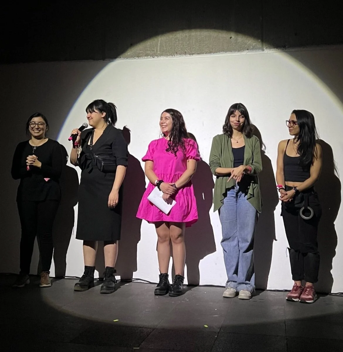 Cinco mujeres de pie bajo un foco de luz, con una de ellas sosteniendo un micrófono.