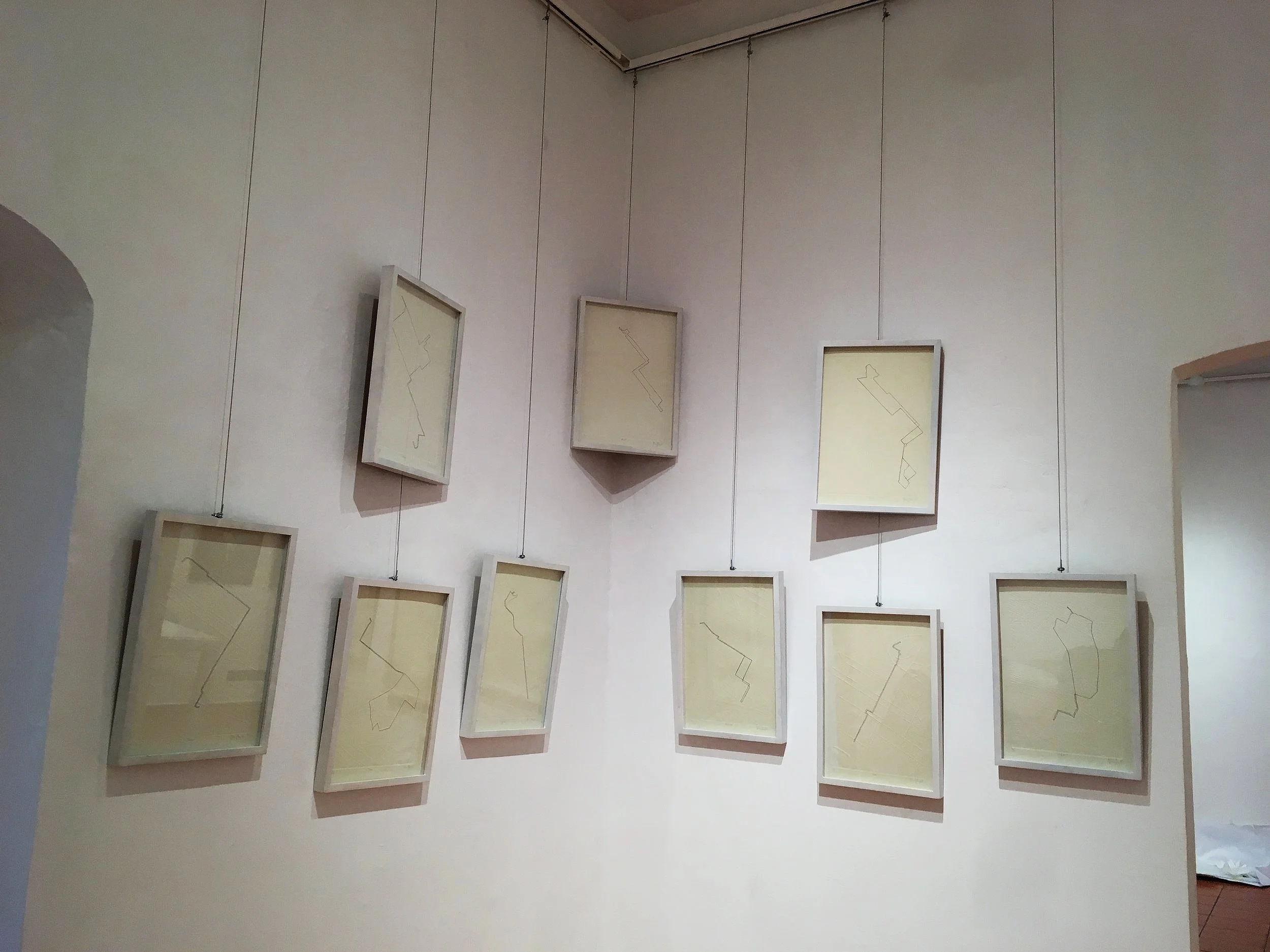 Varios cuadros colgados en una pared, cada uno con un dibujo lineal en un color claro sobre un fondo beige. Los cuadros están sujetos con cables desde el techo.