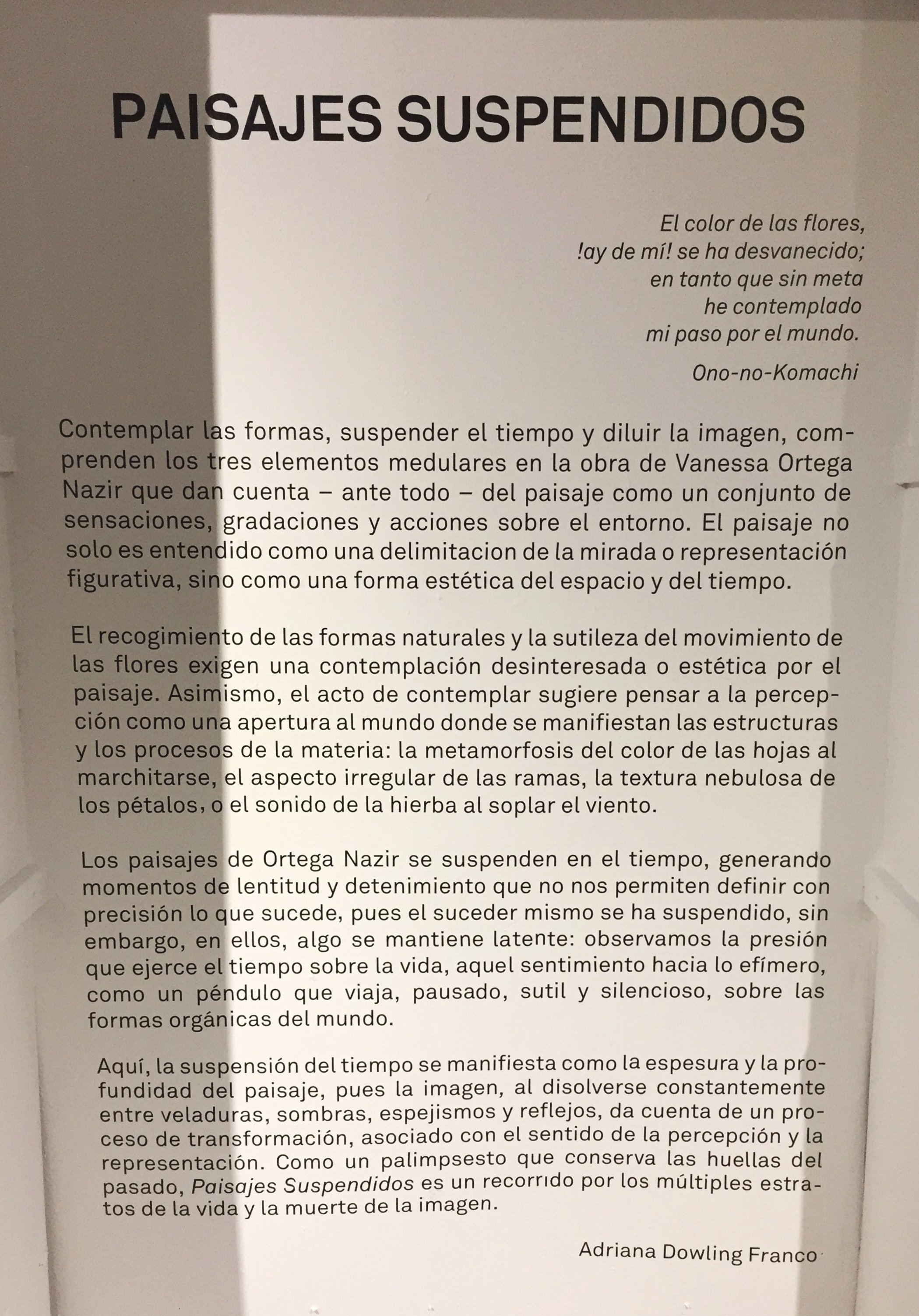 Texto en una pared que describe "Paisajes Suspendidos" por Vanessa Ortega Nazir, mencionando elementos como la contemplación de formas, suspensión del tiempo y disolución de imágenes. La obra se centra en estructuras naturales y procesos de transformación, creando un sentido de tiempo pausado y sutil. Incluye una cita de Ono-no-Komachi y es escrito por Adriana Dowling Franco.