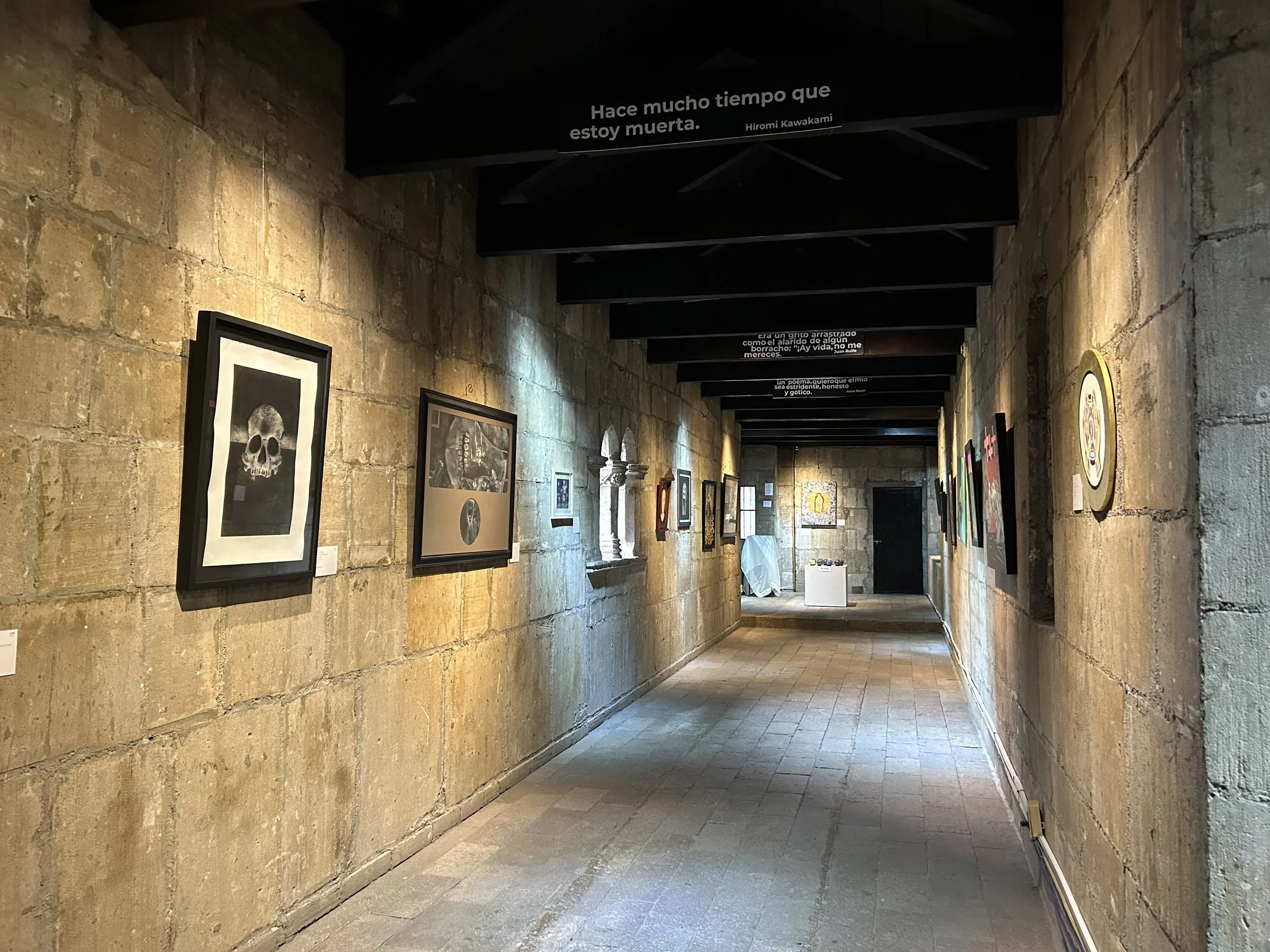 Pasillo de una exposición de arte con paredes de piedra, cuadros colgados y citas escritas en el techo.