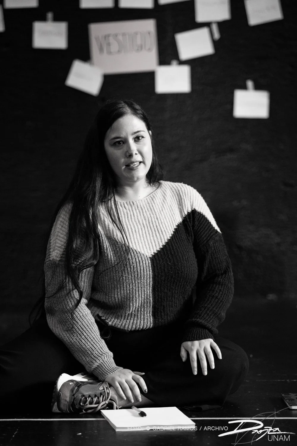 Mujer sentada en el suelo con un suéter de colores oscuros, hablando o presentando, con una hoja de papel y bolígrafo frente a ella. Detrás hay papeles colgados en la pared.