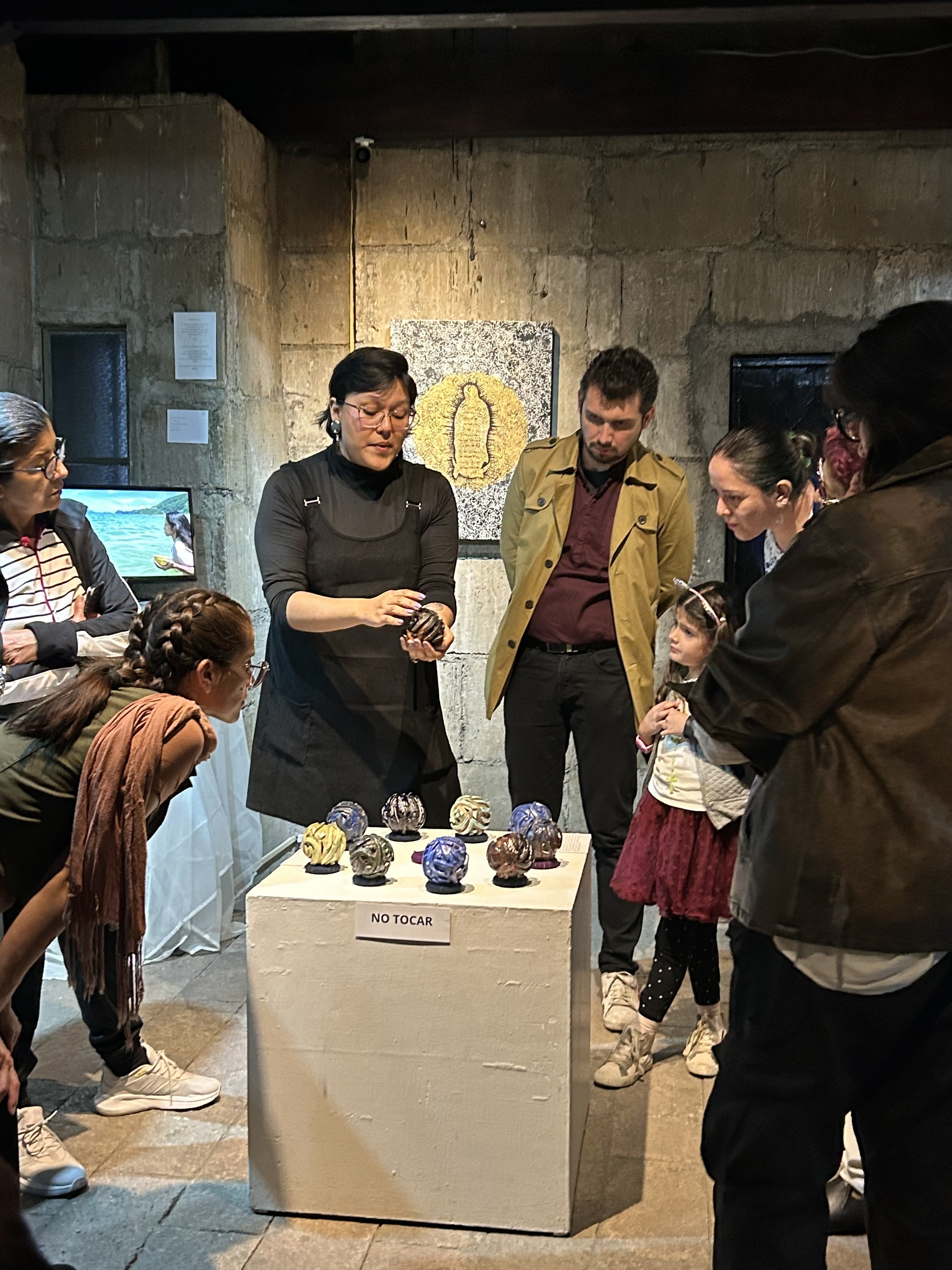 Un grupo de personas observando una presentación de arte en una galería. En el centro, hay una persona mostrando un objeto pequeño. Sobre una mesa, hay varias esculturas pequeñas de colores. Un cartel indica "NO TOCAR".