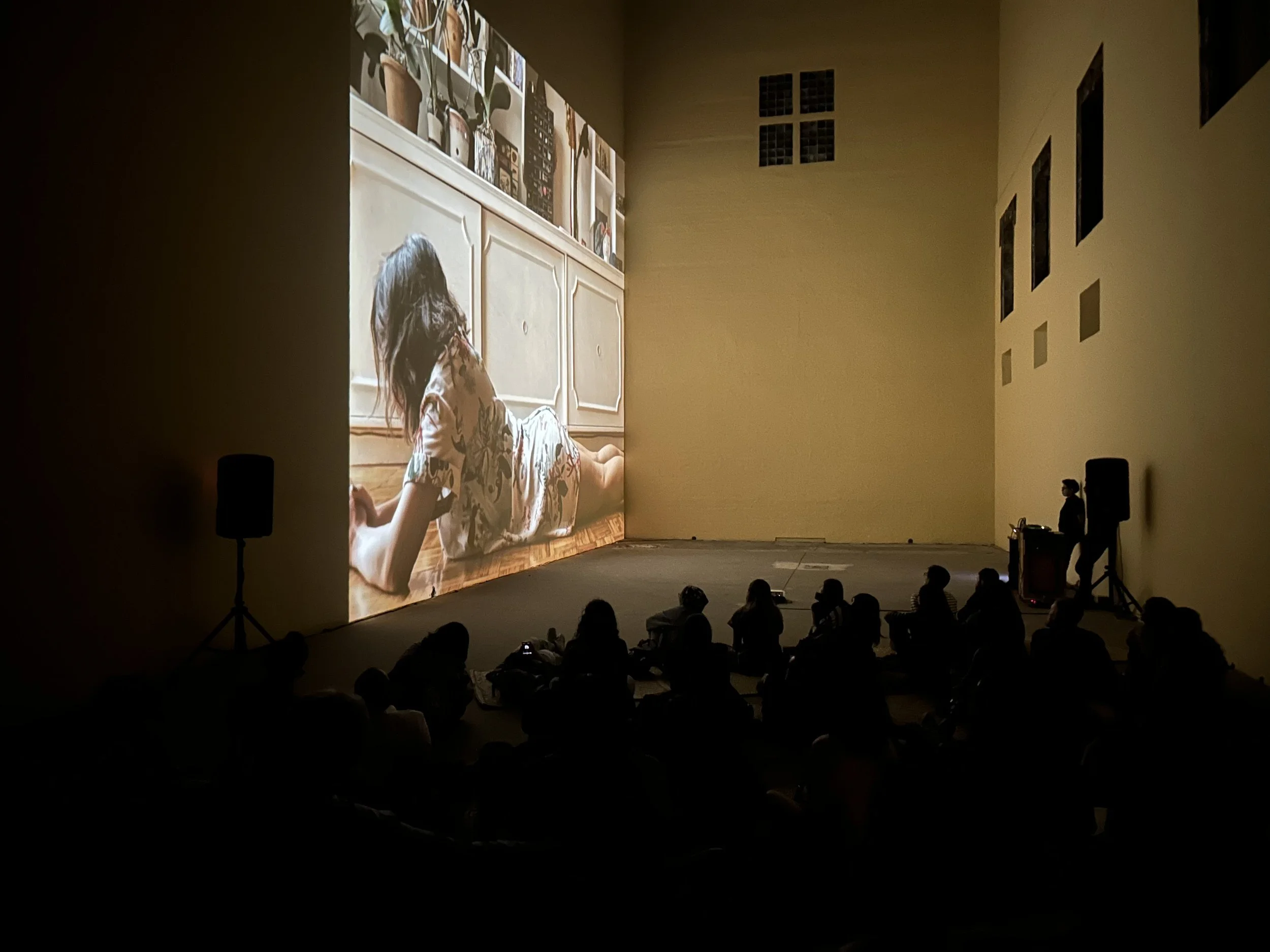 Sala oscura con personas viendo una proyección de video mostrando a una persona acostada en el suelo frente a un mueble blanco.