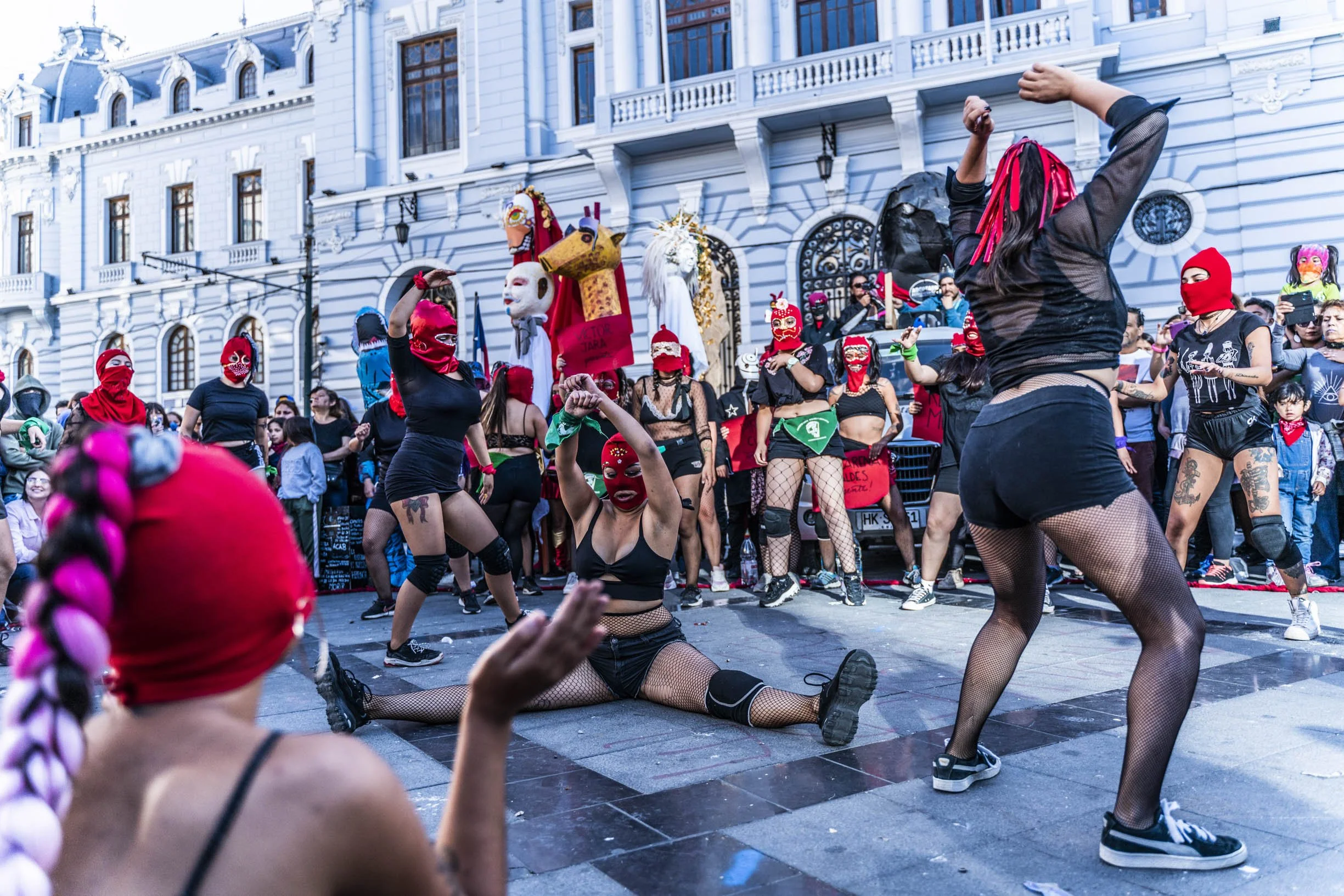 Personas enmascaradas realizando una performance en una calle frente a un edificio histórico. Llevan ropa negra y algunas usan medias de red. Hay espectadores observando el evento.