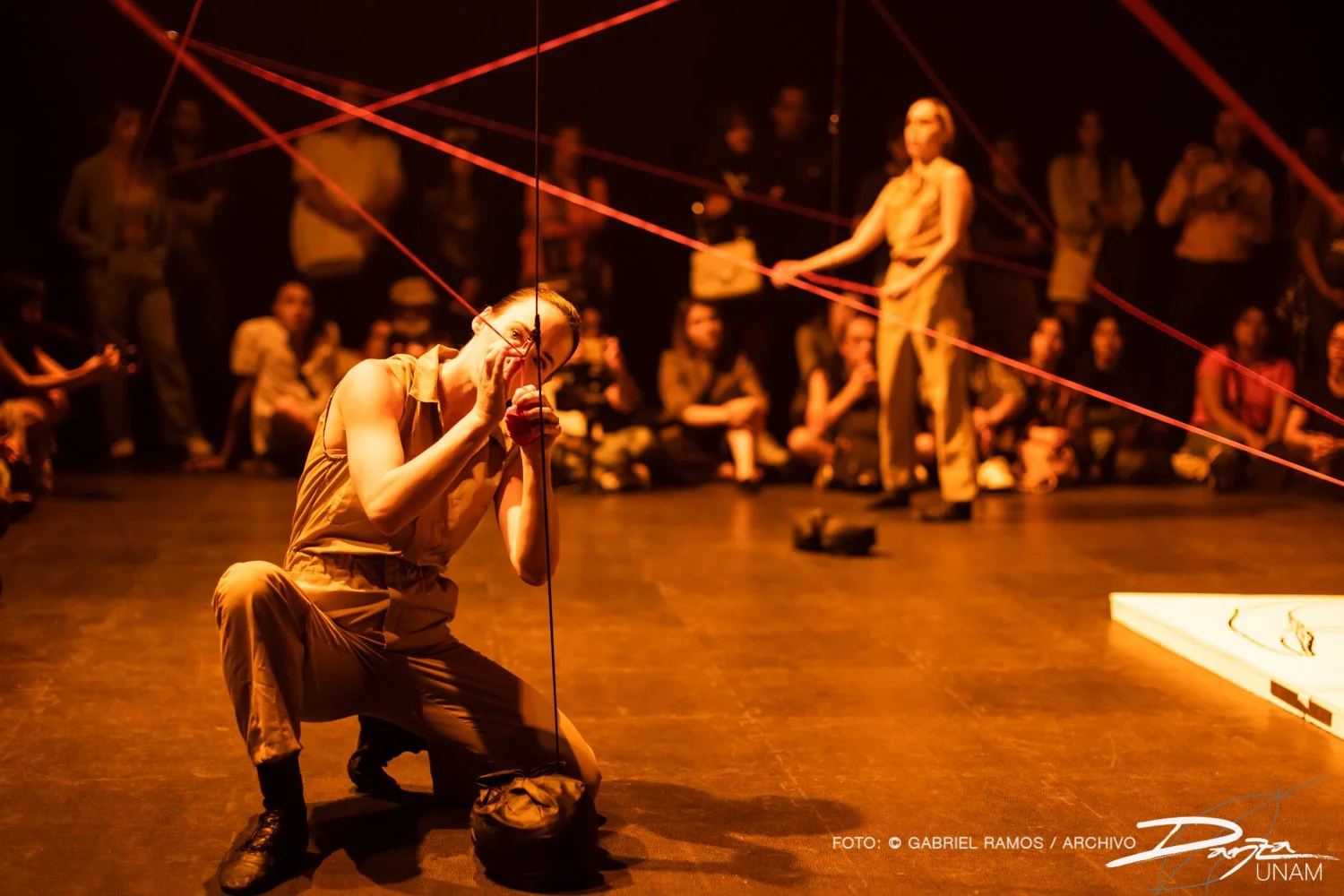 Escena teatral con una persona actuando, arrodillada, sosteniendo cuerdas rojas. Fondo con espectadores observando. Ambiente de iluminación cálida y artística.