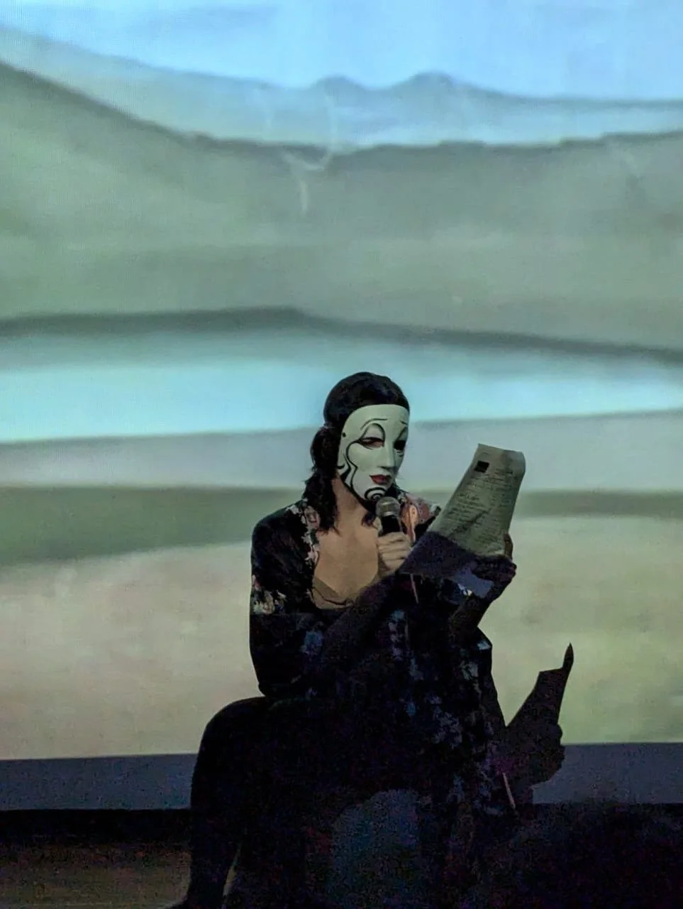 Persona con máscara blanca pintada sosteniendo un micrófono y leyendo un papel, sentada frente a una proyección de paisaje.