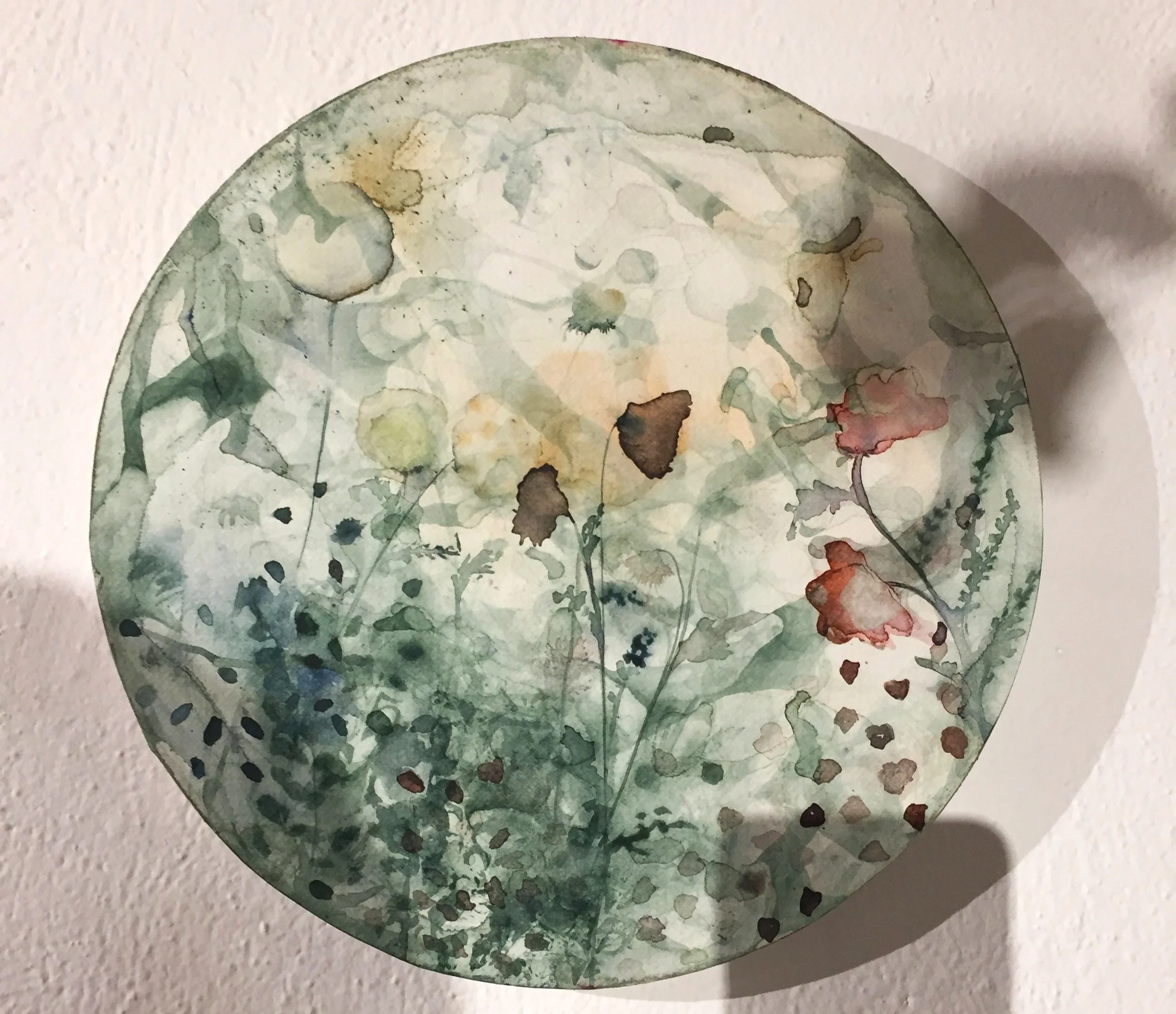 Pintura circular en acuarela con flores y hojas en tonos verdes, amarillos y rojos, sobre un fondo blanco.