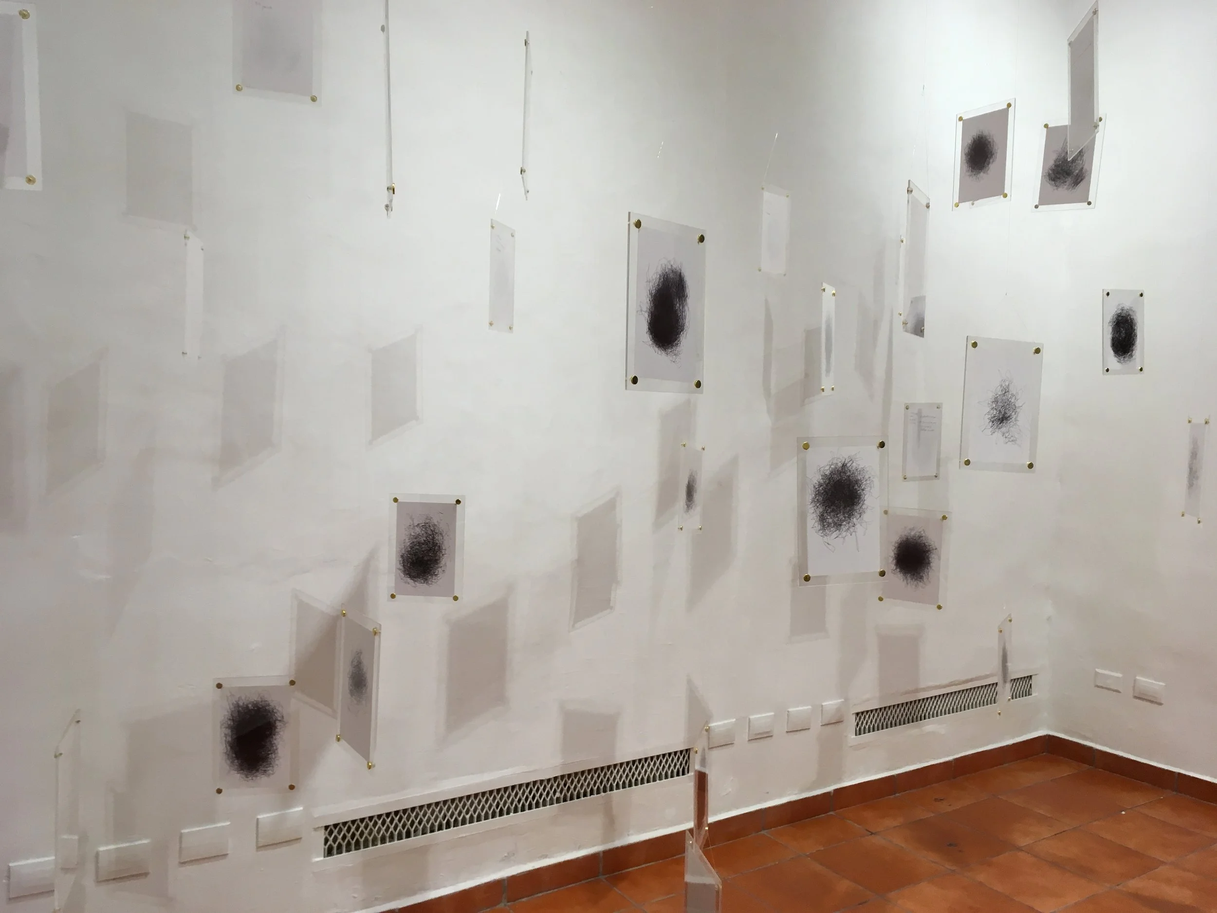 Instalación artística en una galería con dibujos abstractos enmarcados, colgados en una pared blanca, pisos de baldosas.