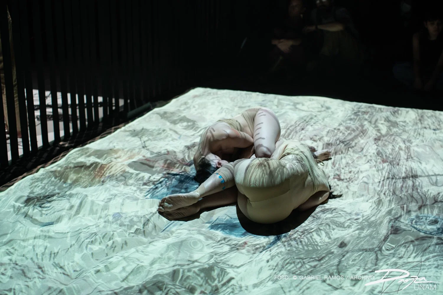 Dos personas en trajes de cuerpo completo de color beige, acurrucadas en el suelo iluminado con proyecciones artísticas.