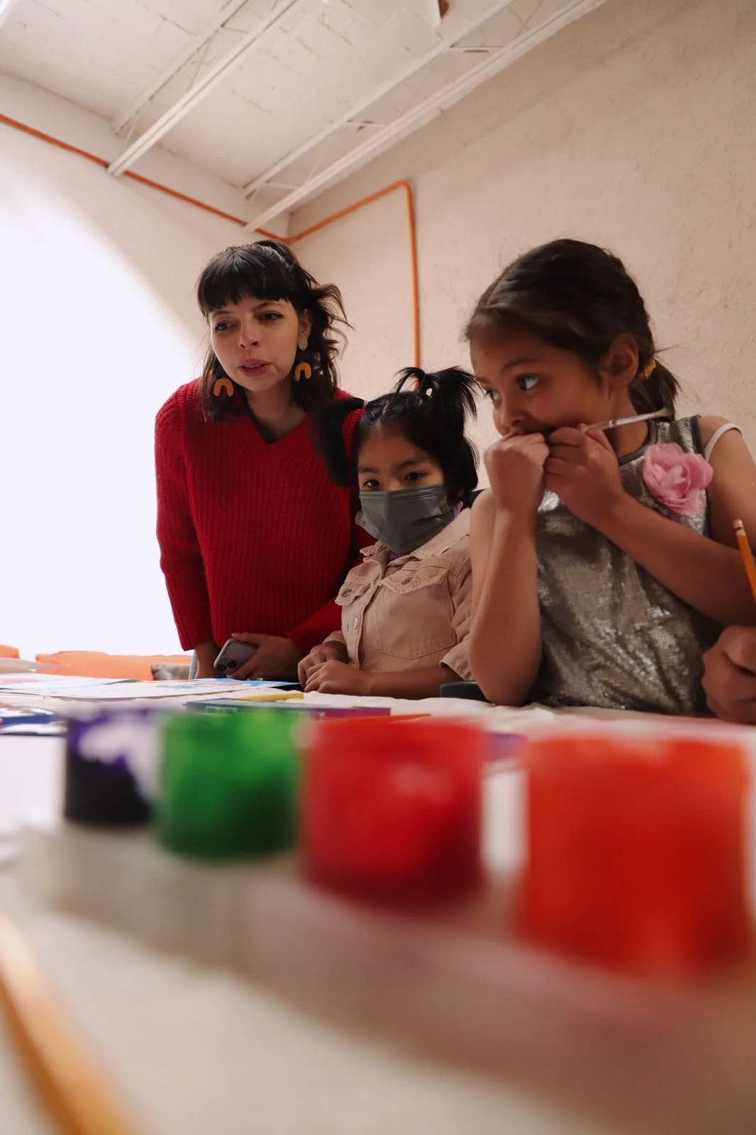 Un grupo de personas en una mesa blanca con frascos de pintura y pinceles visibles en primer plano. Detrás, hay un adulto explicando algo a dos niños, uno de ellos usando mascarilla.