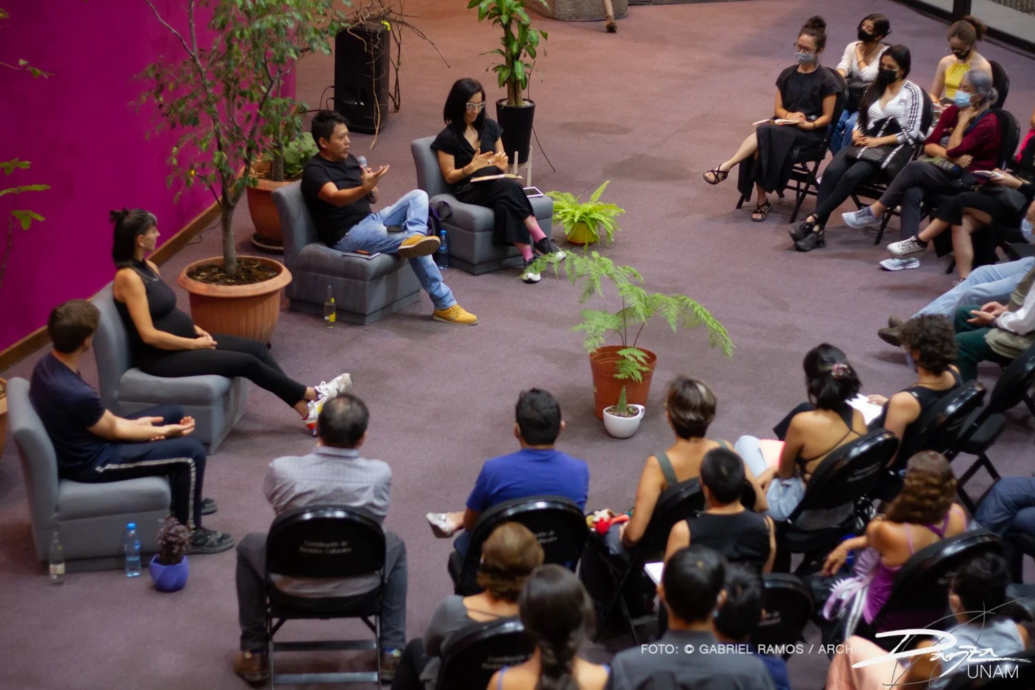 Personas sentadas en círculo participando en una charla o discusión en un espacio interior con plantas de interior y decoración colorida.