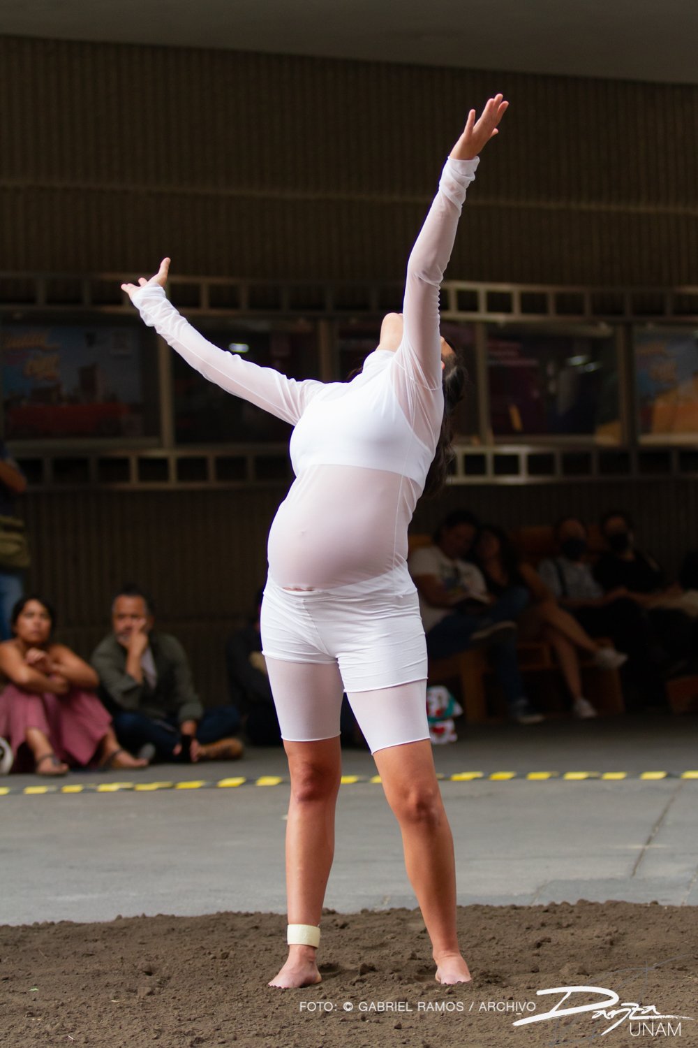 Mujer embarazada vestida de blanco realizando una actuación de danza en un espacio público, con público observando.