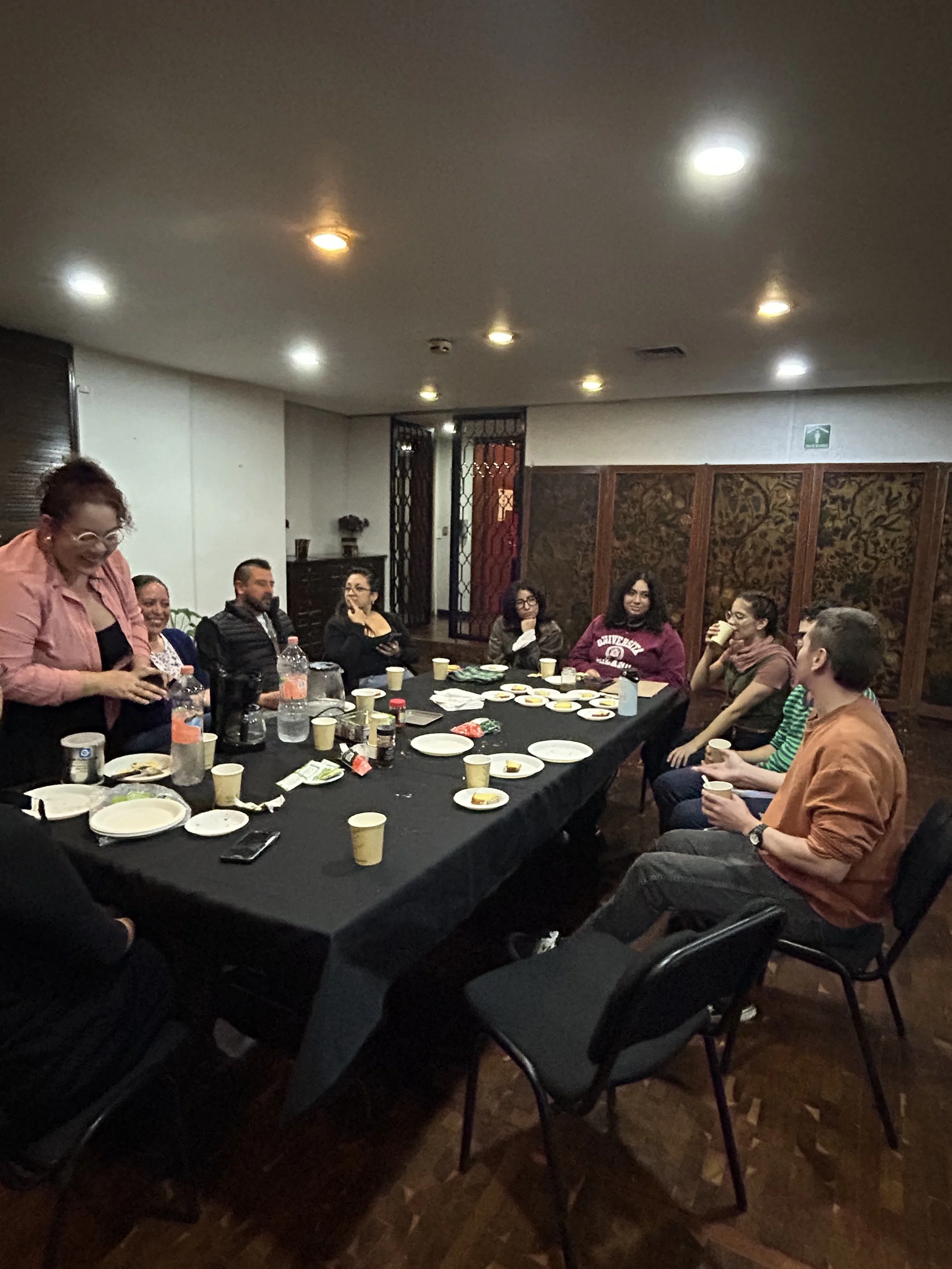 Personas reunidas alrededor de una mesa con comida y bebidas, en una sala iluminada.