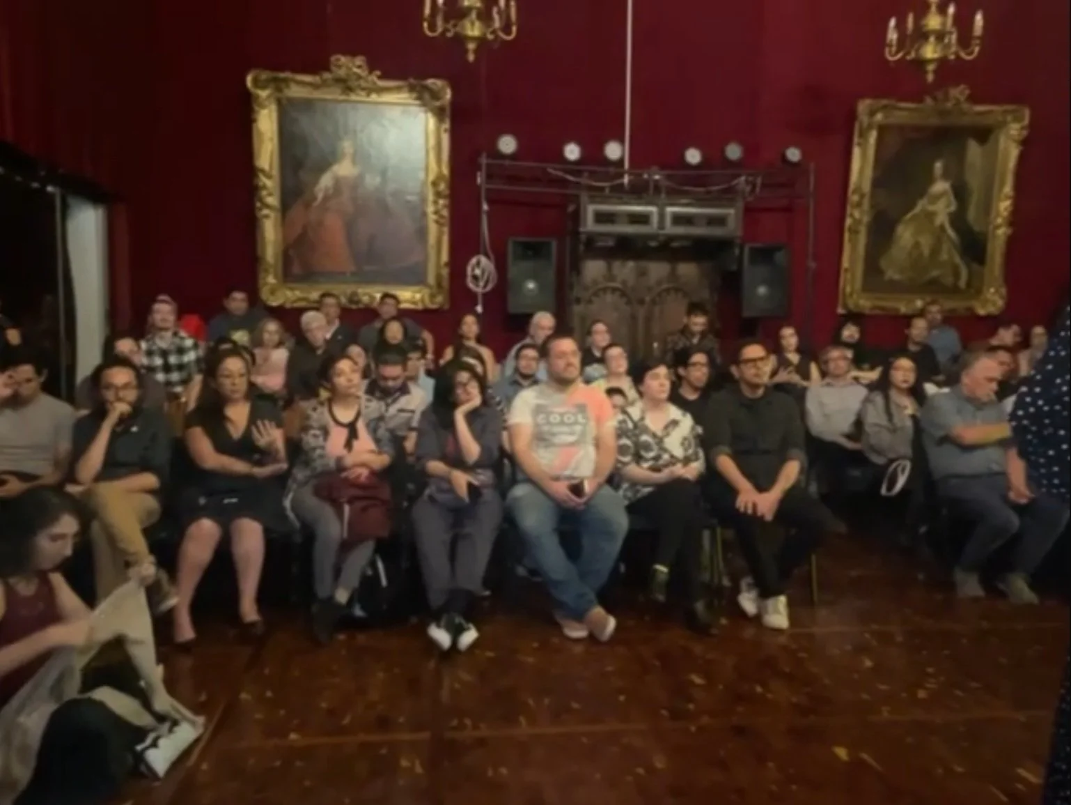 Grupo de personas sentadas en un auditorio con paredes rojas y cuadros grandes enmarcados, observando hacia el frente.