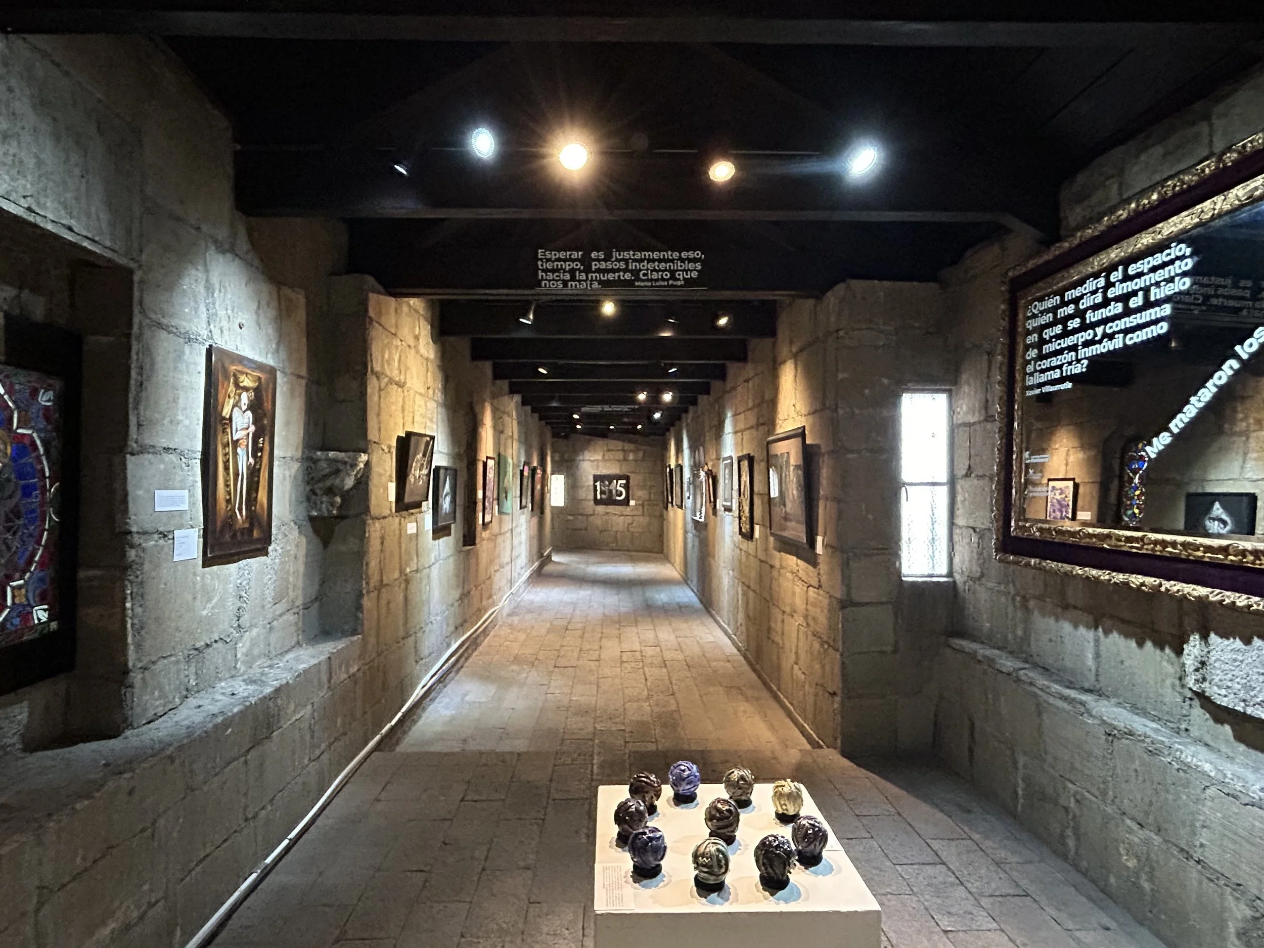 Pasillo de galería de arte con cuadros en las paredes y esculturas en una mesa central, iluminación tenue y decoración de piedra.