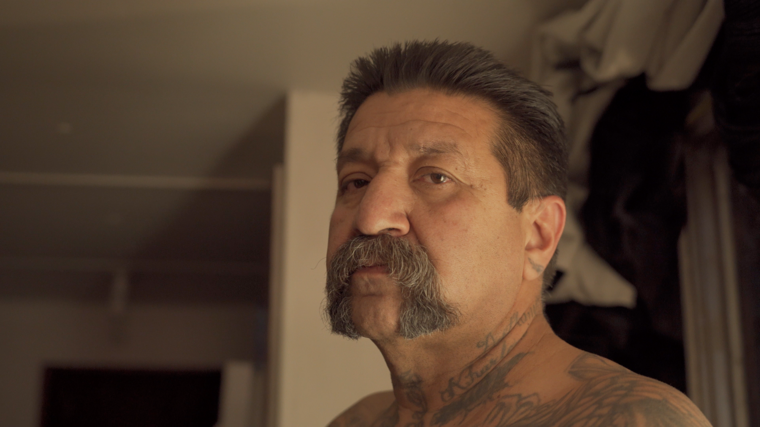 Hombre con bigote y tatuajes mirando hacia la cámara en una habitación.