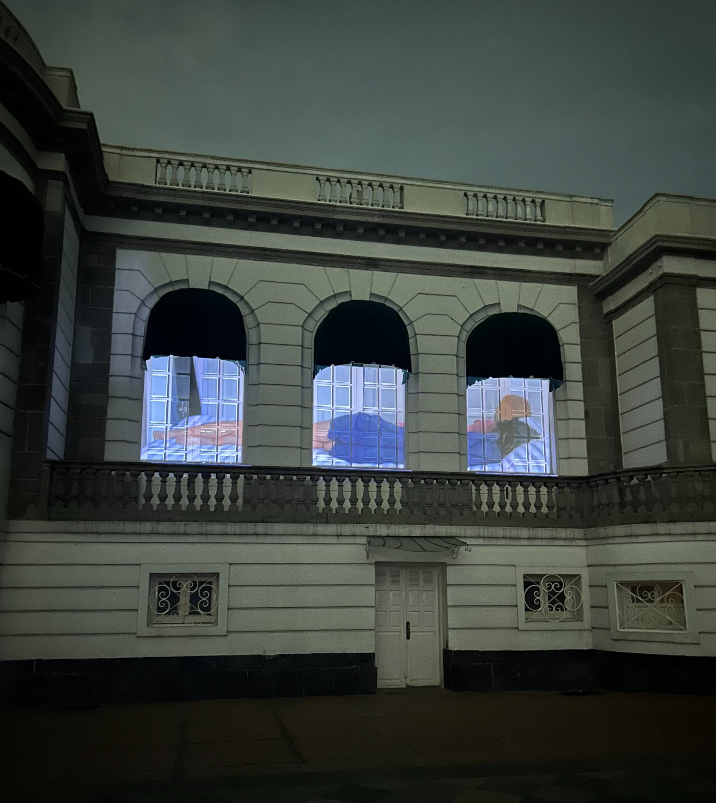 Fachada de edificio clásico con ventanas iluminadas desde adentro, mostrando imágenes o proyecciones. Arquitectura de estilo antiguo con arcos y ornamentación.
