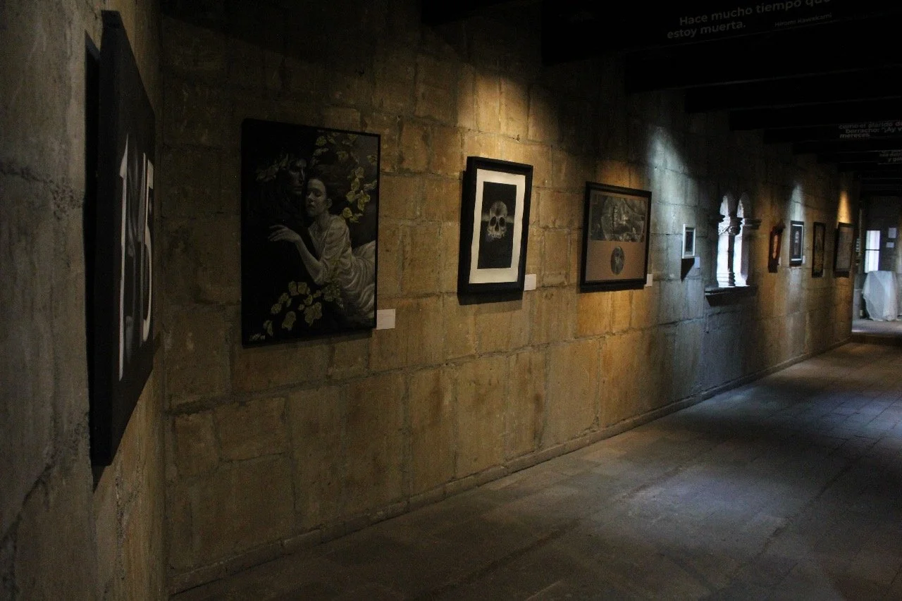 Pasillo de galería de arte con pinturas y dibujos colgados en la pared de piedra, algunas obras enmarcadas y una ventana pequeña.