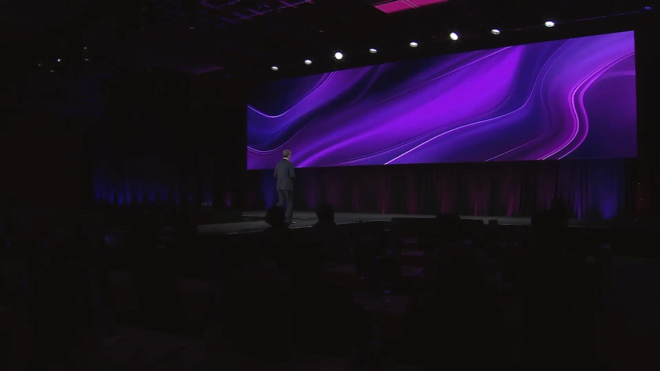 A person on stage at a conference or presentation, facing a large screen with swirling purple abstract graphics.