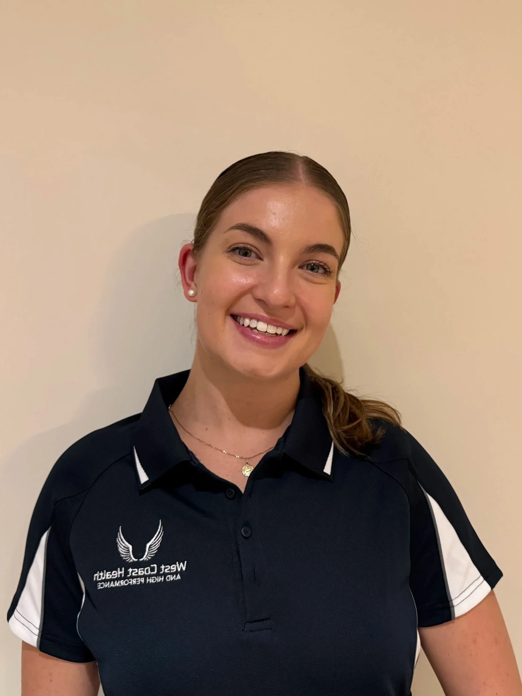 A smiling woman with brown hair tied back, wearing a navy blue polo shirt with white accents, standing against a beige wall.