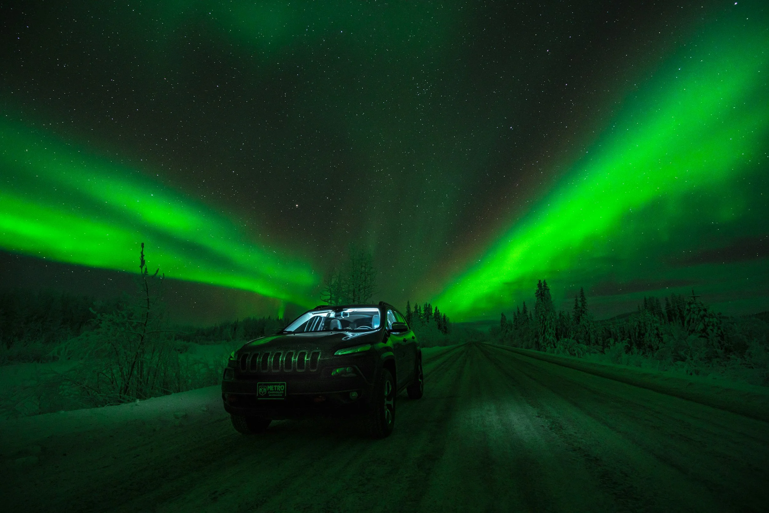 A black SUV parked on a snow-covered road at night, with green northern lights illuminating the sky and stars visible in the background.