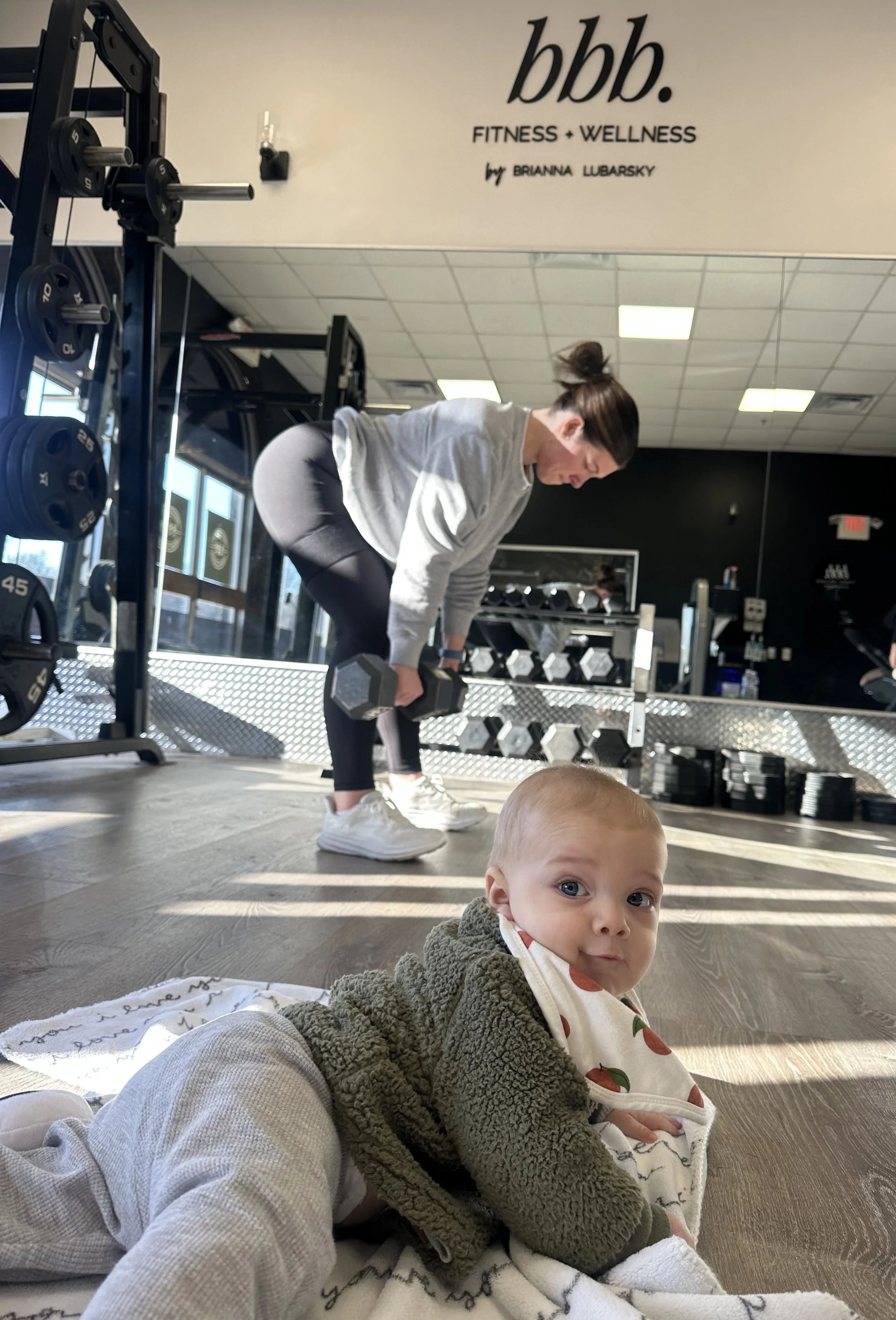 A woman is lifting a dumbbell in a gym, while a baby is lying on the floor in the foreground. The gym has weights and mirrors, with a sign on the wall that reads 'bbb. FITNESS + WELLNESS by Brianna Lubarsky'.