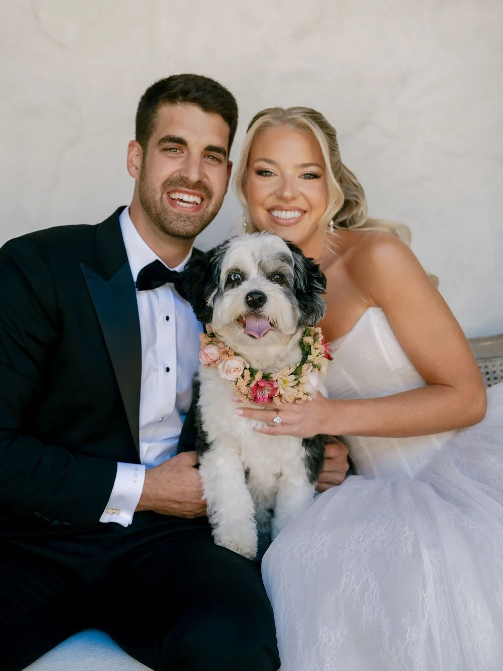 Bride and groom smiling with their black and white mini Aussie doodle wearing a pink and yellow floral collar on their wedding day.