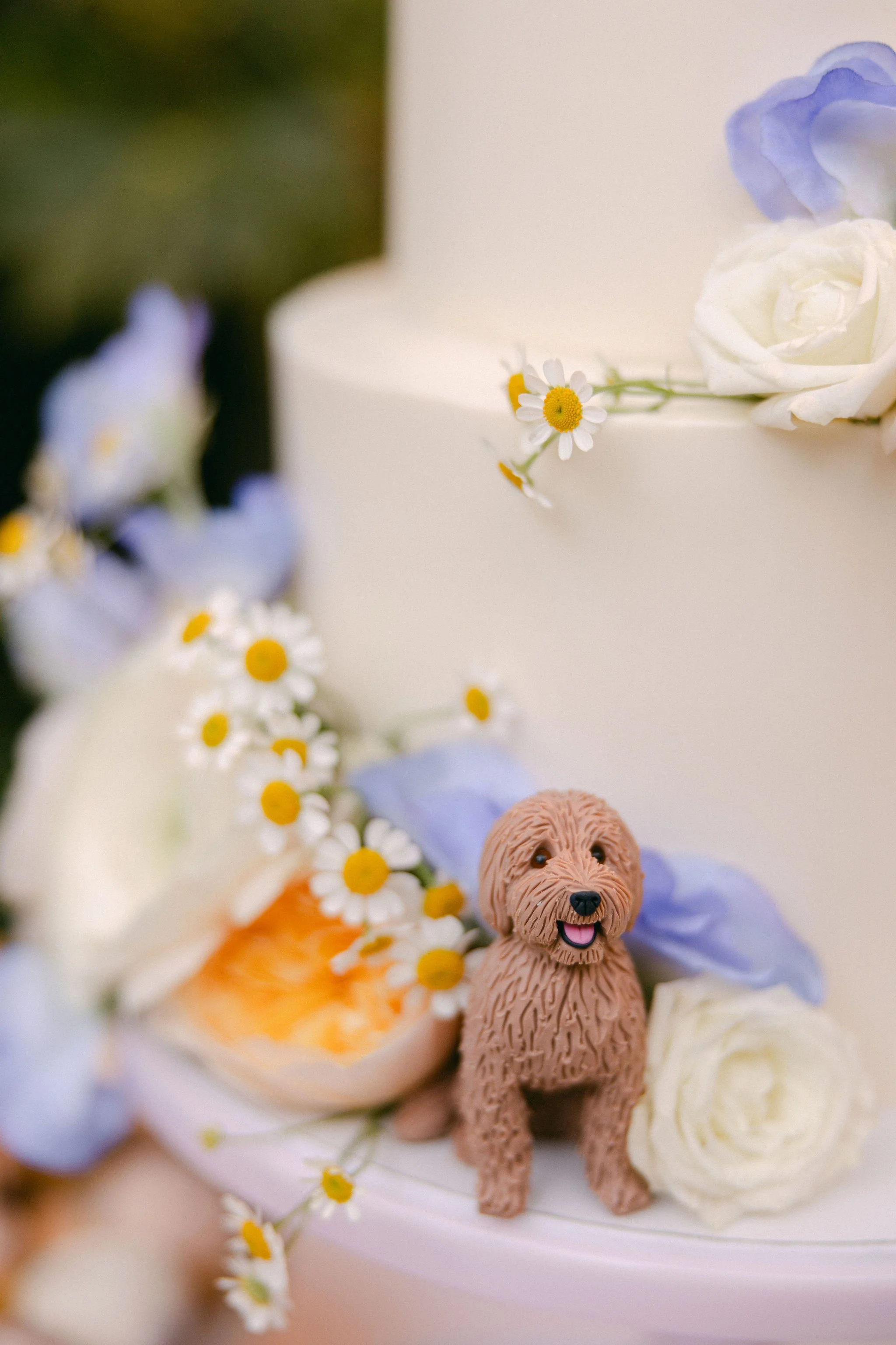 A close-up of a white wedding cake’s lower tier, showing a small brown dog cake topper nestled among peach roses, white ranunculus, daisies, and lavender petals.