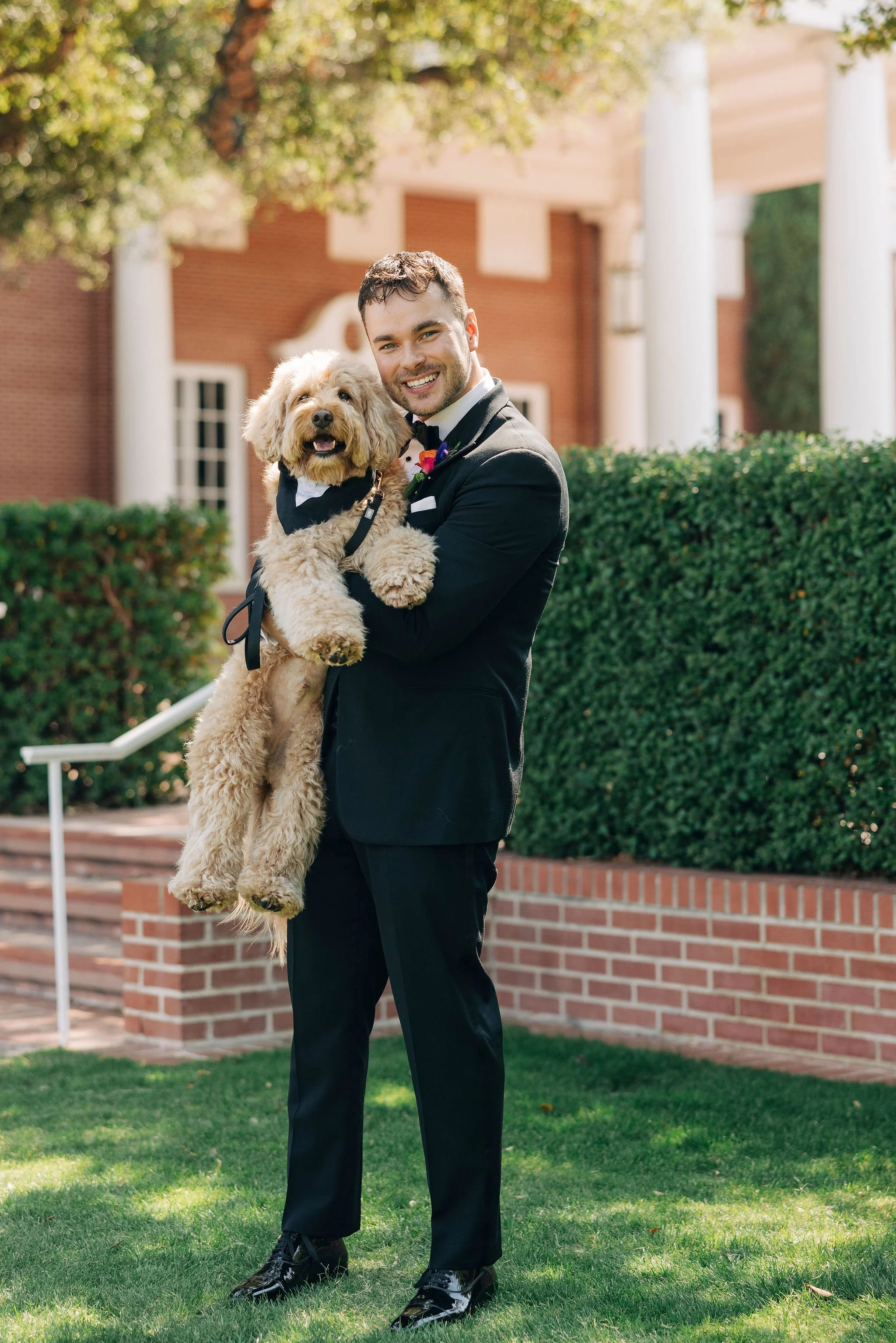 A man in a black tuxedo holding a dog wearing a tuxedo outdoors in front of a red brick building with white columns and green bushes.