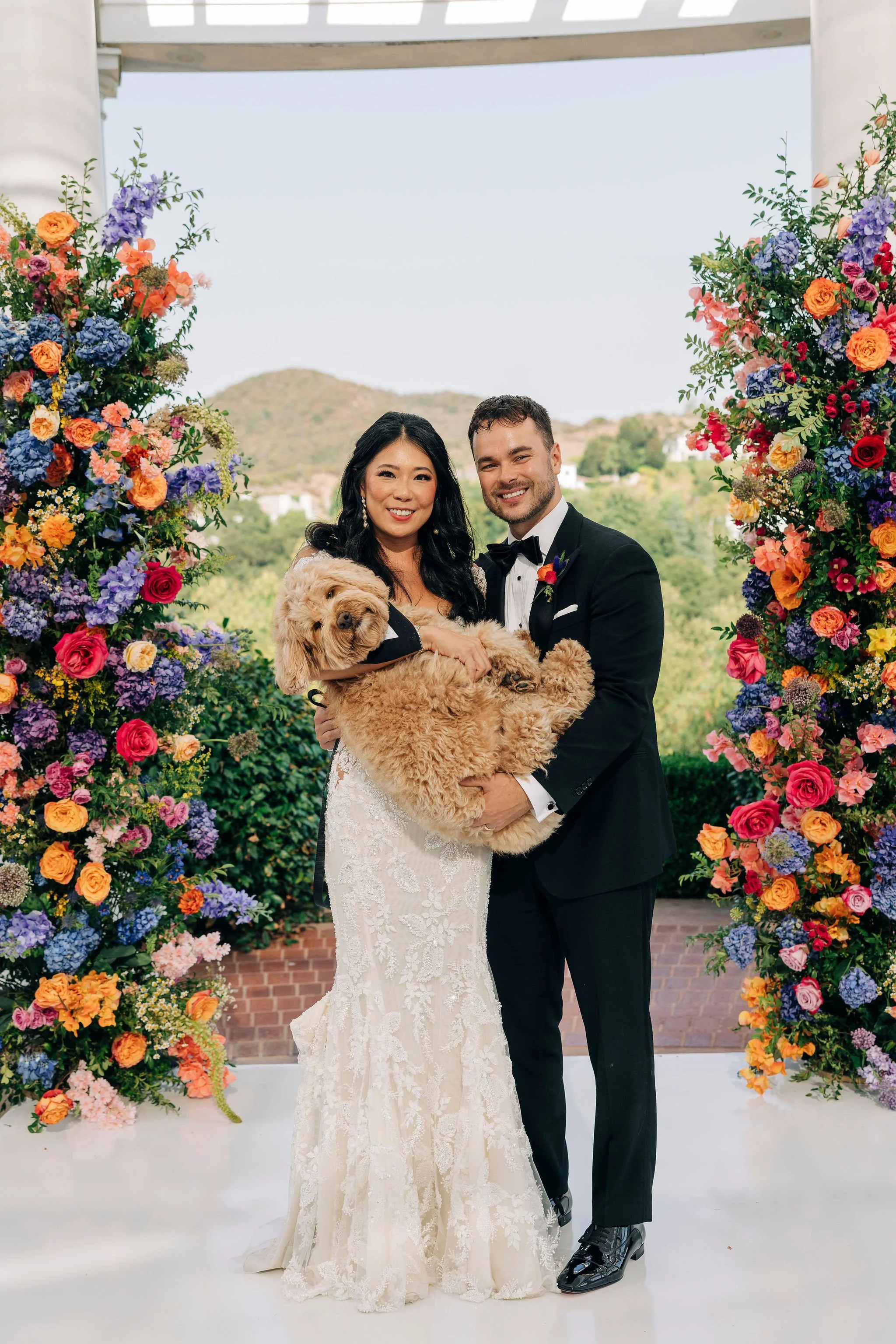 A newlywed couple, dressed in wedding attire, poses outdoors with their dog in front of a colorful floral arch. The bride holds the dog, which is wearing a bow tie, and the couple is smiling at the camera with a scenic hillside background.