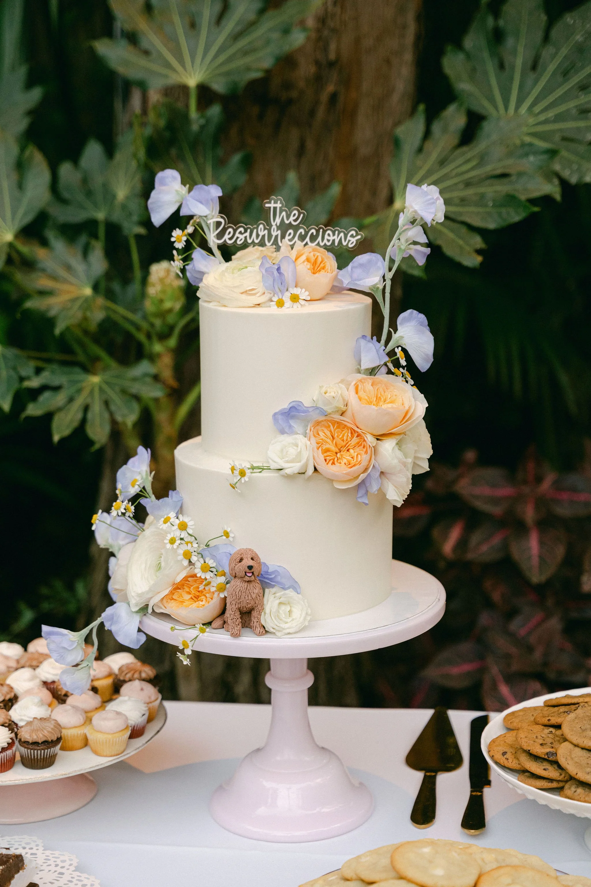 A two-tier white wedding cake is decorated with pastel flowers in peach, white, and lavender, and a small figurine of a curly-haired dog sits at the base of the cake.There is a custom cake topper reading ‘The Resurreccions’ placed on top.