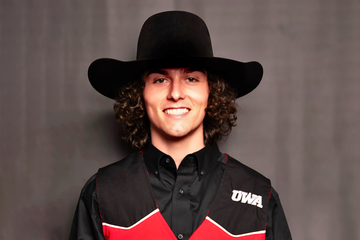 A man with curly hair, wearing a black wide-bramed cowboy hat, black shirt, and a black vest with red and white accents, smiling at the camera against a grey background.