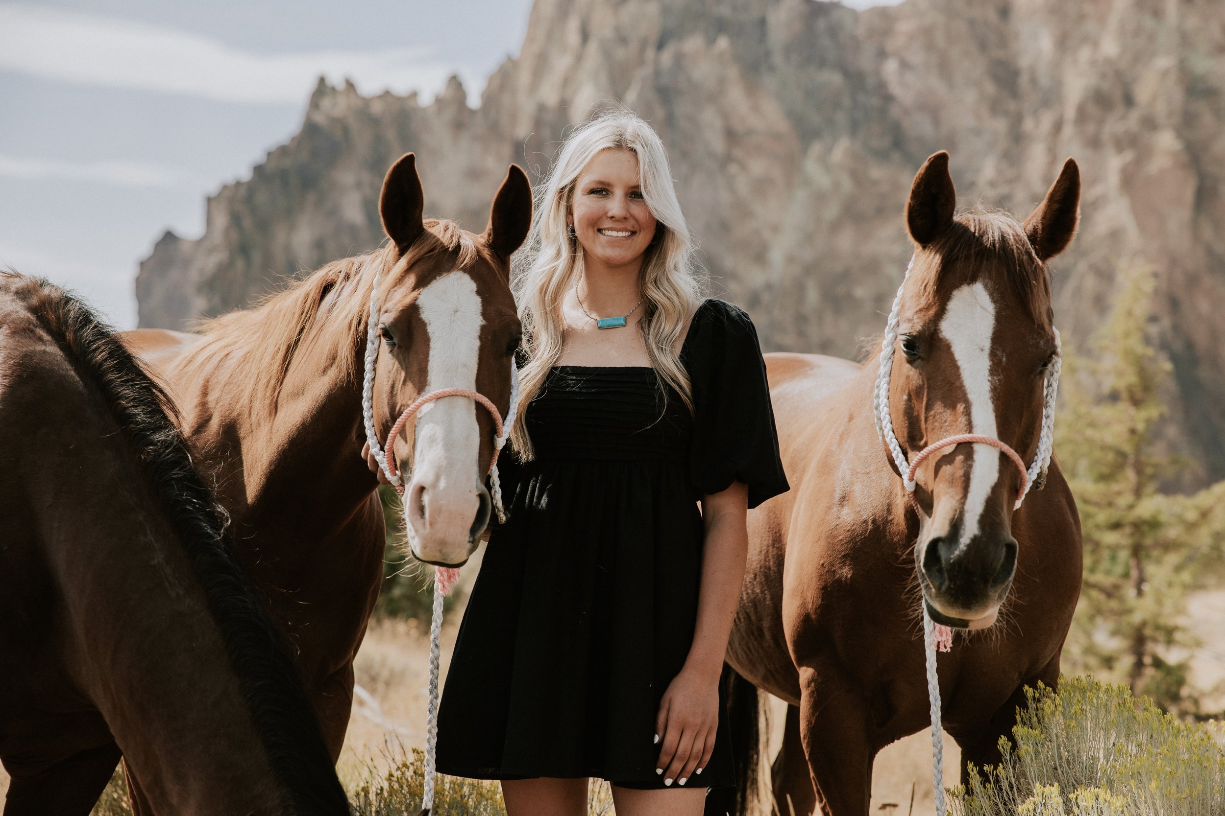 A woman with long blonde hair in a black dress standing between two brown horses with white markings, outdoors in a grassy area with mountains in the background.
