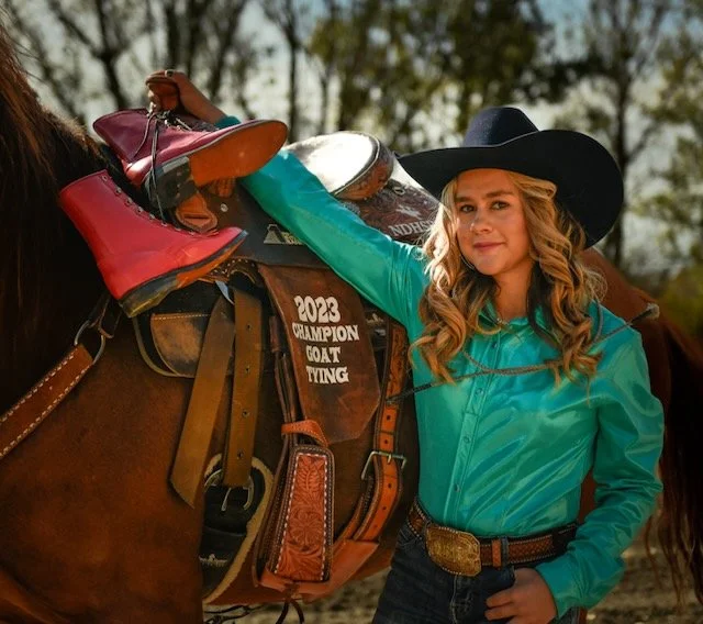 Young woman in western attire with a cowboy hat standing next to a horse, adjusting its saddle decorated with colorful boots and a sign that says '2013 Champion Goat Tying'.