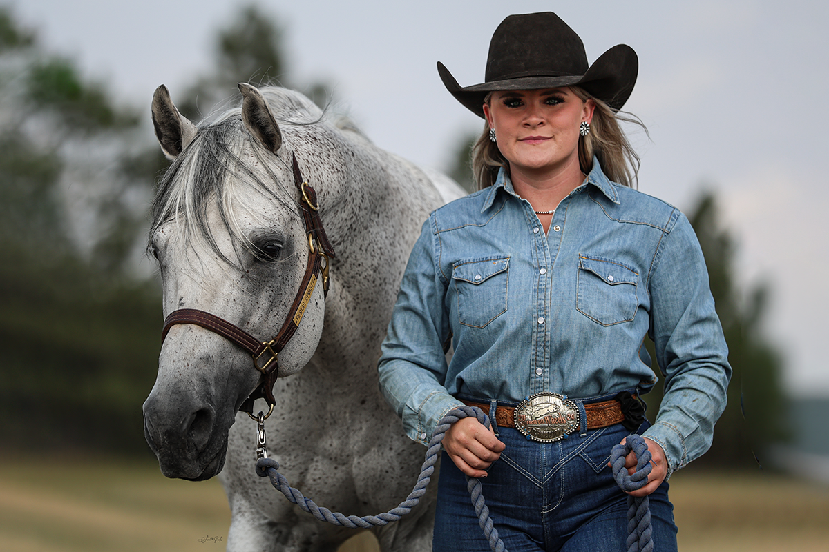 A woman in a cowboy hat, denim shirt, and jeans standing with a gray speckled horse outdoors.