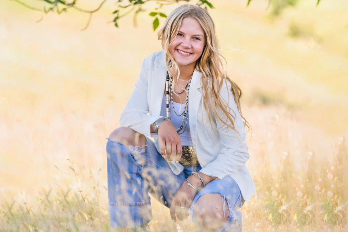 Young woman with long blonde hair smiling while kneeling in a field of tall grass with green leaves and trees in the background, wearing a white jacket, distressed jeans, and multiple jewelry accessories.