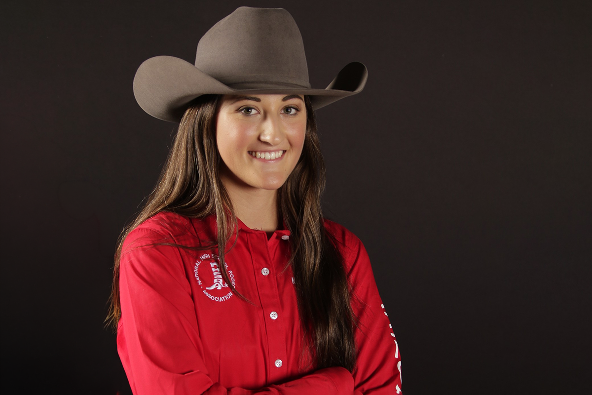 A young woman wearing a large cowboy hat and a red shirt with a high school logo, standing against a dark background.