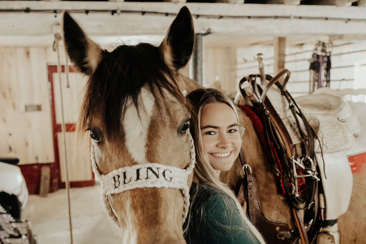 A smiling young woman standing next to a brown horse with a white stripe on its face, inside a wooden barn with riding equipment.