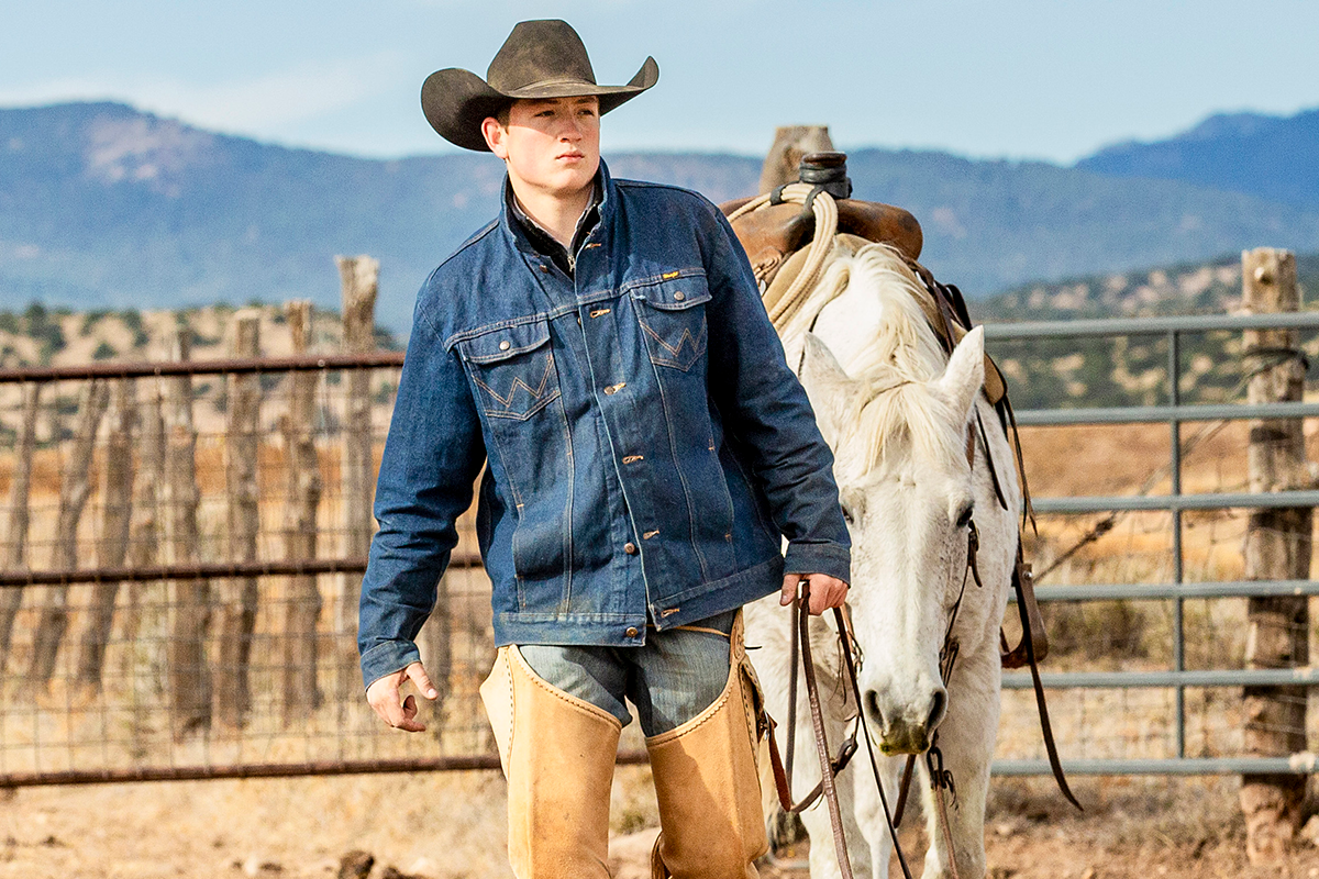 A young man dressed as a cowboy standing next to a white horse on a ranch.