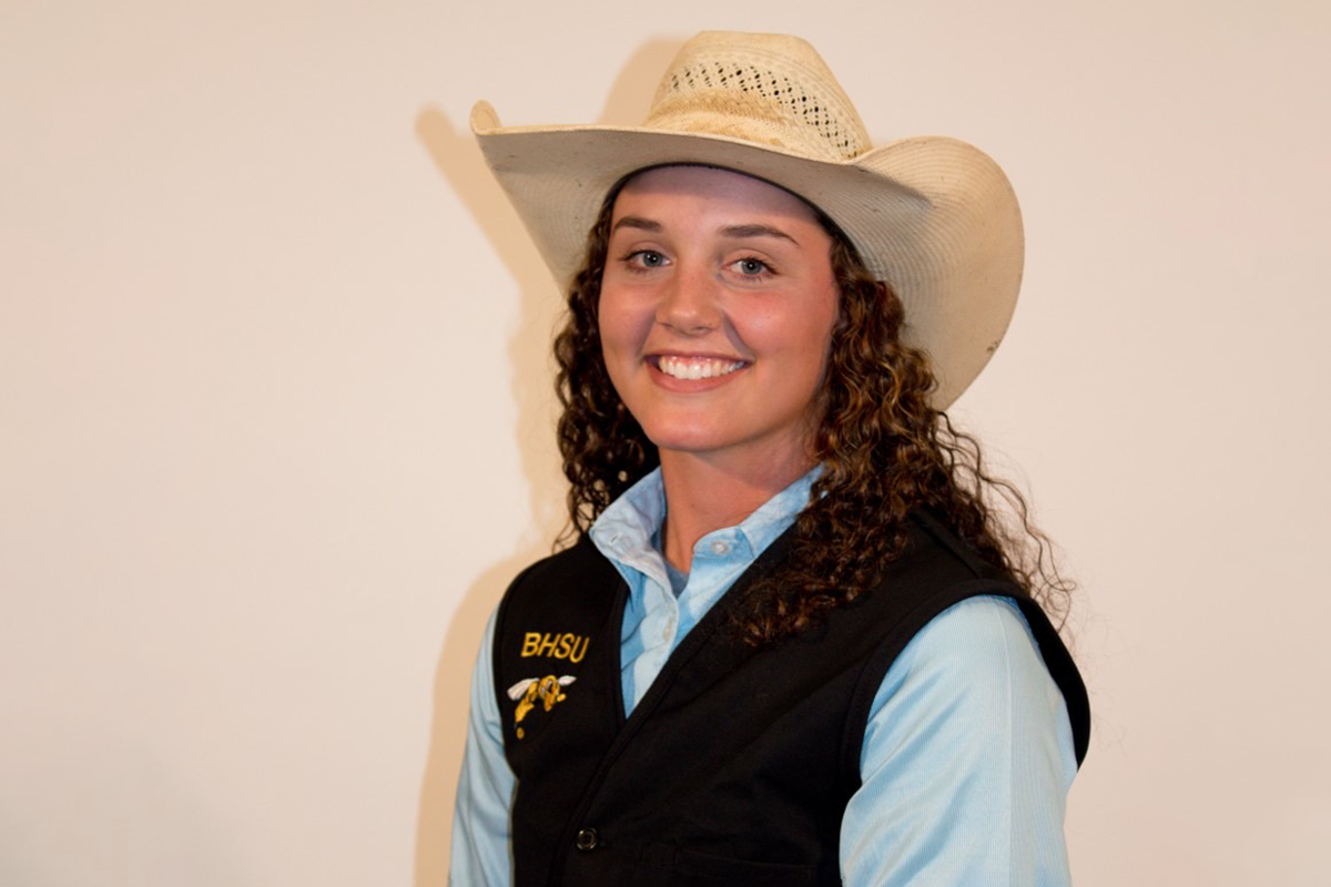 Young woman with curly hair smiling, wearing a cowboy hat, a light blue button-up shirt, and a black vest with the letters "BHSU" and a yellow bee logo.