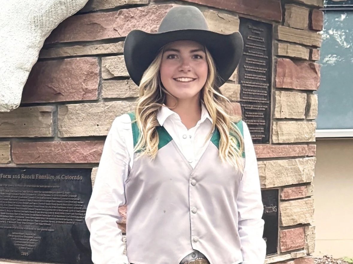 Young woman dressed as a park ranger, wearing a gray cowboy hat, white shirt, and beige vest, standing in front of a brick wall.