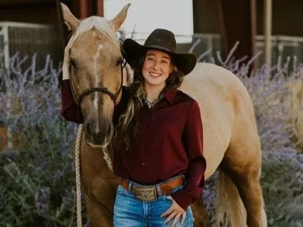 A young woman with long hair wearing a black hat, maroon shirt, and jeans standing next to a tan and white horse.