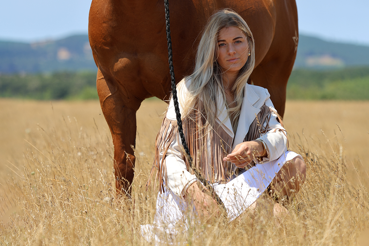 A young woman with blonde hair sitting on the grass in a field, next to a brown horse, with hills in the background and clear sky.
