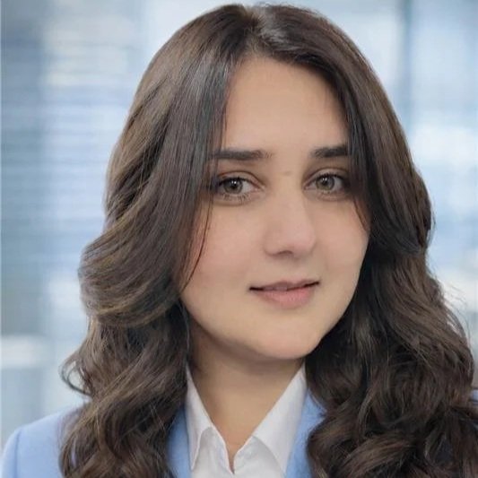 A woman with long, wavy brown hair wearing a white blouse and light blue blazer, standing indoors with a cityscape background observed through large windows.