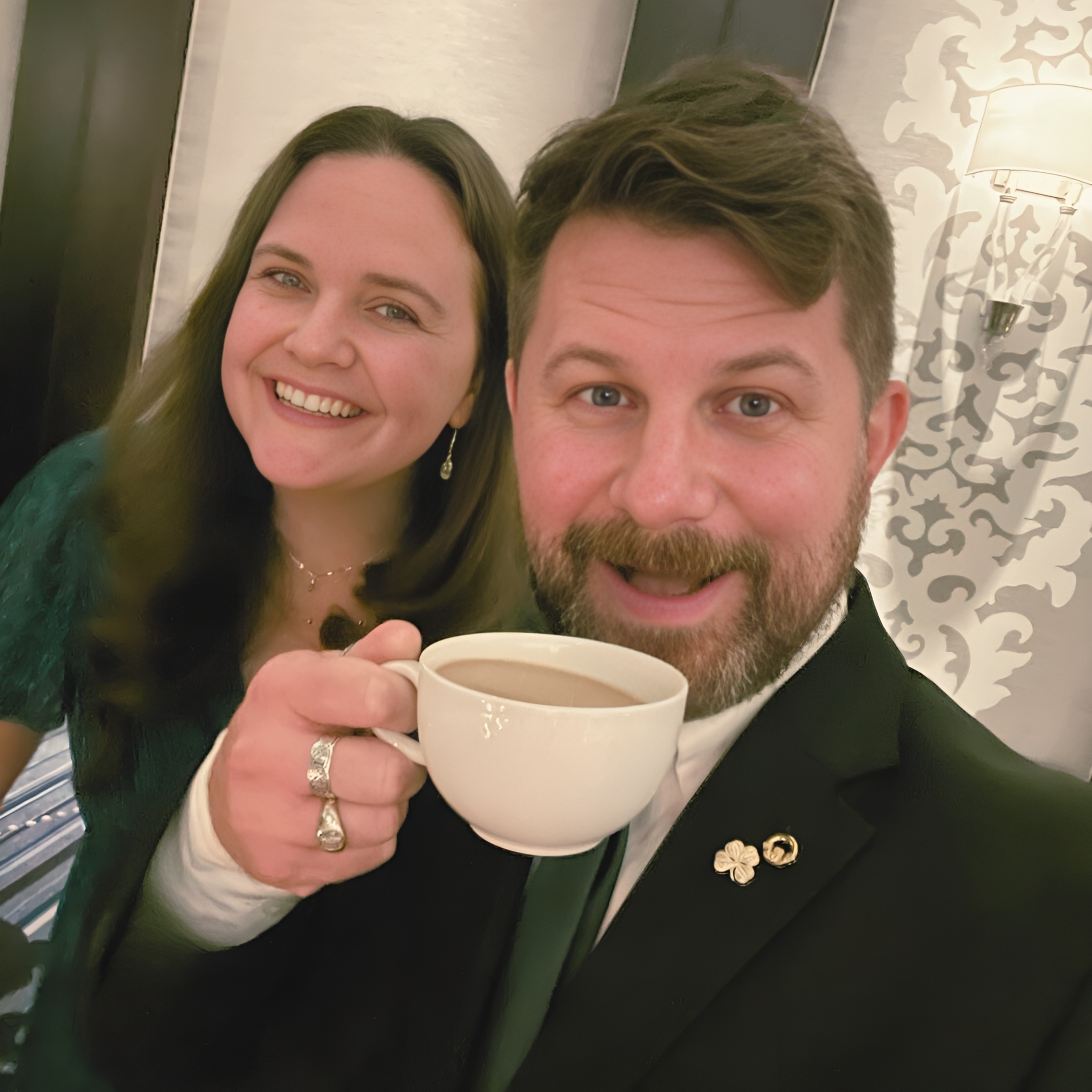 A smiling woman and man dressed formally, holding a cup of coffee, in an elegantly decorated room.