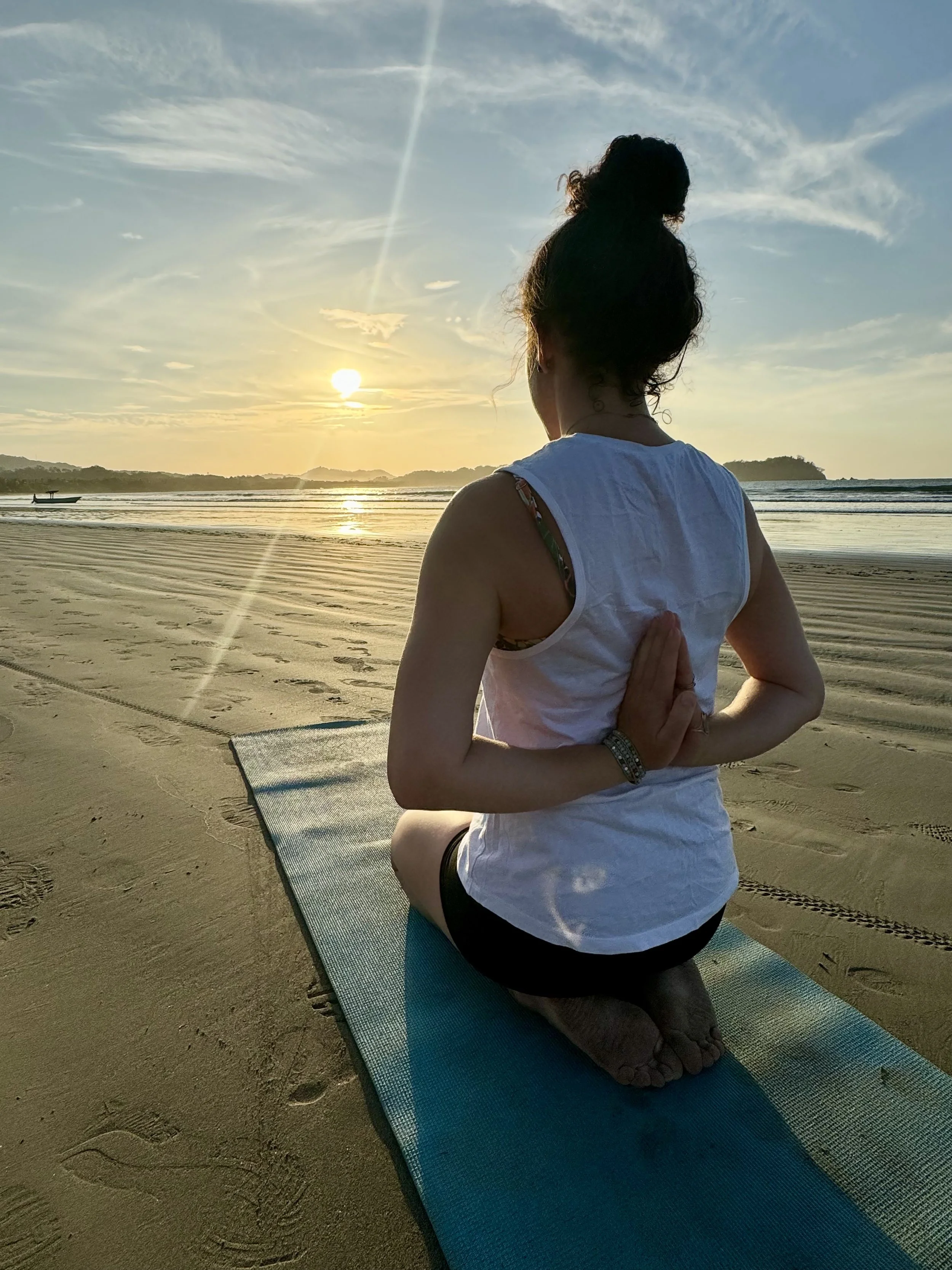 A woman practicing yoga on a beach during sunset, sitting on a yoga mat with her back to the camera, in a prayer position behind her back.