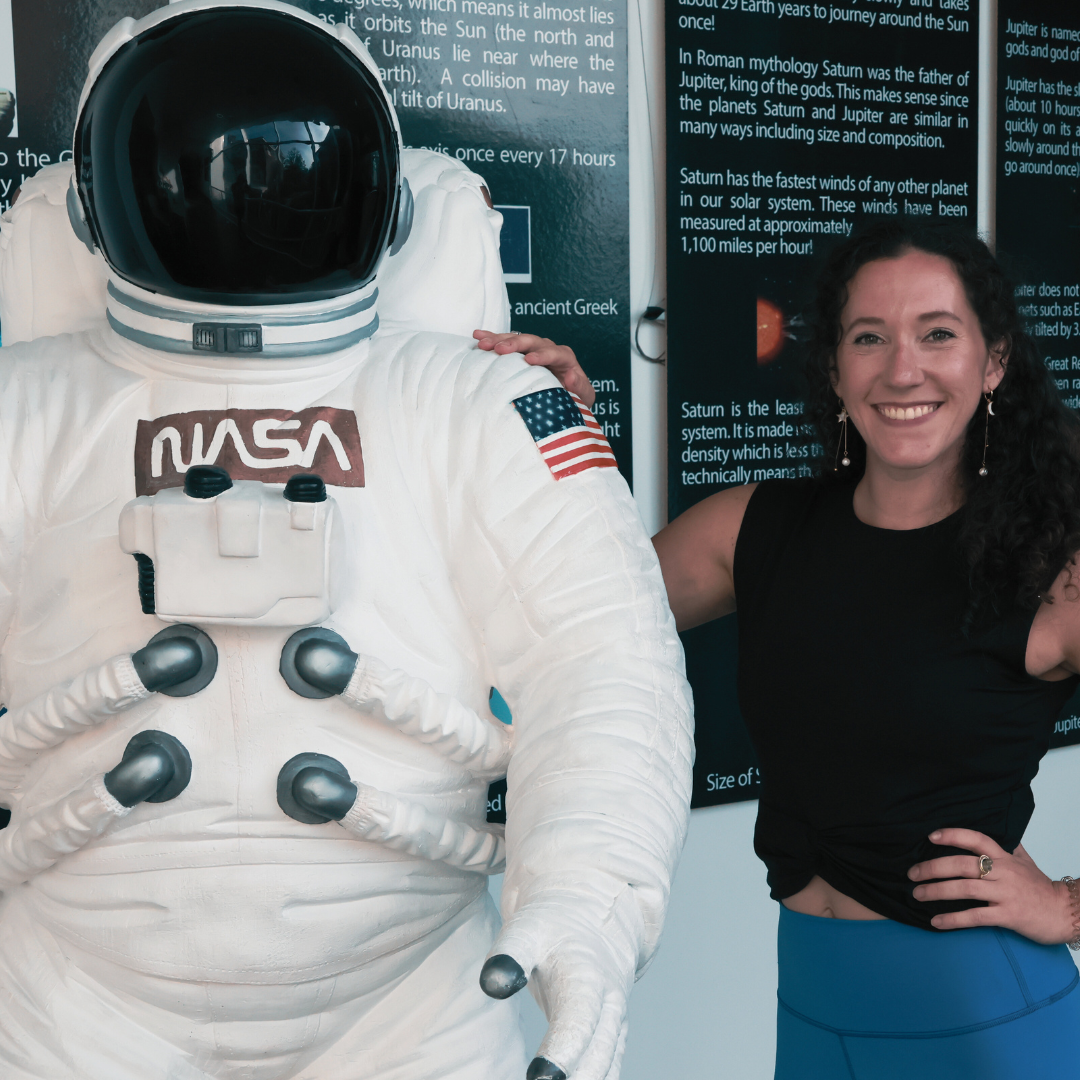 A woman smiles with her arm around an astronaut mannequin at a space exhibit. The mannequin wears a white NASA spacesuit with an American flag patch on the shoulder and a black reflective helmet. There are informational posters about planets in the background.