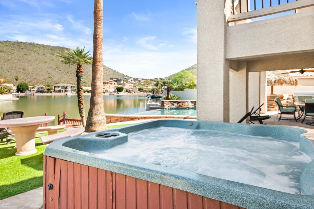 Outdoor hot tub with bubbling water on a patio with lawn, palm trees, and lake view with houses and hills in the background.